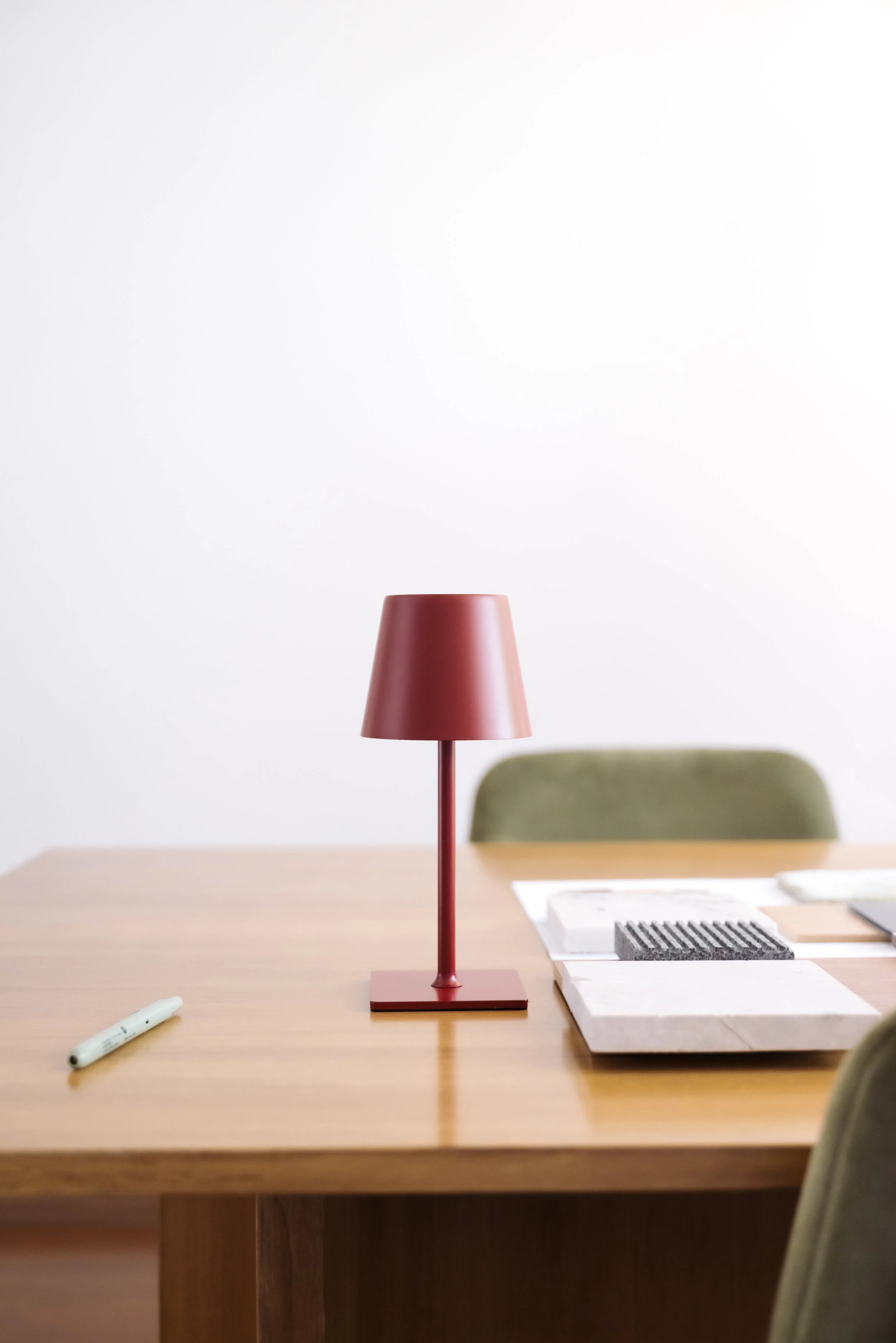 Red table lamp on wooden table with green chair and various papers and objects in a minimalist room.