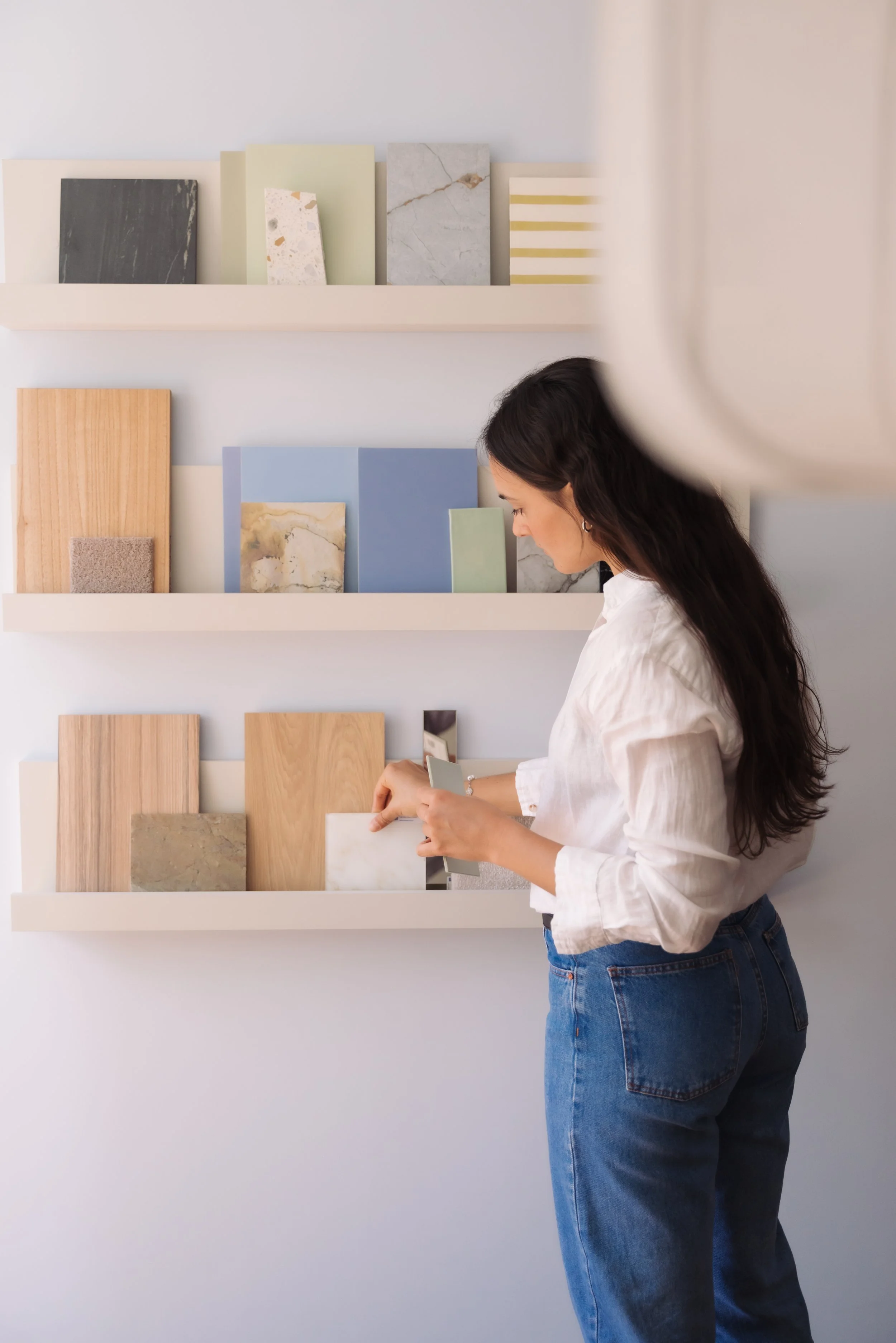 A woman in a white shirt and blue jeans stands in front of wall-mounted shelves displaying various samples of tiles and materials, examining a sample in her hand.