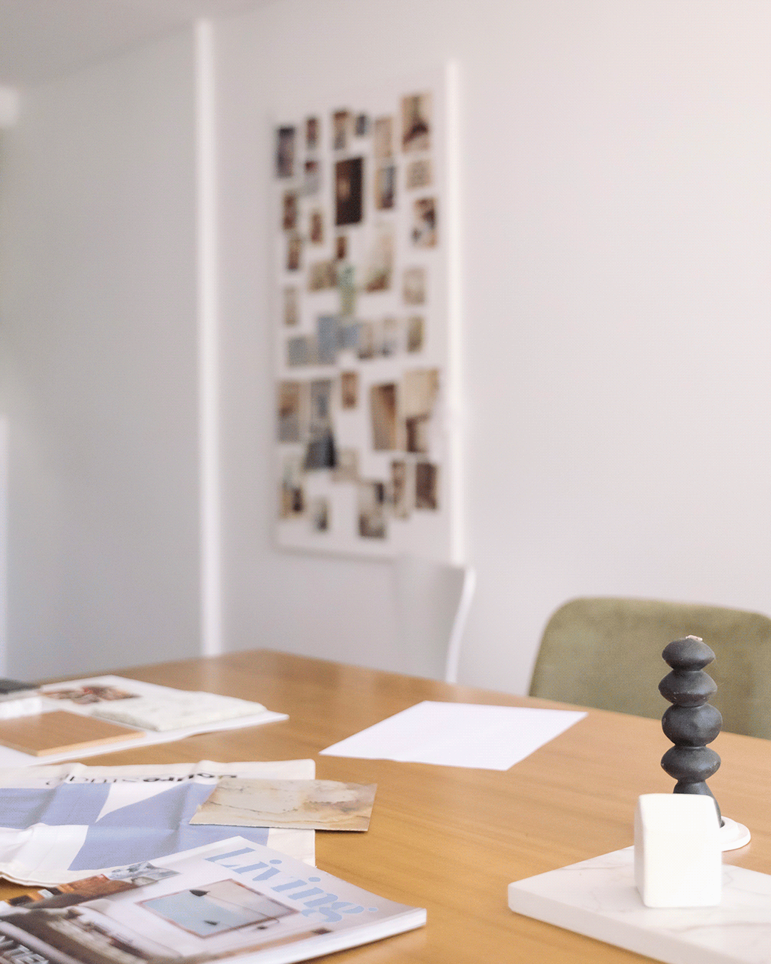 An art or design studio with a wooden table, magazines, and papers, and a decorative stone sculpture on a white stand; blurred collage poster on the wall in the background.