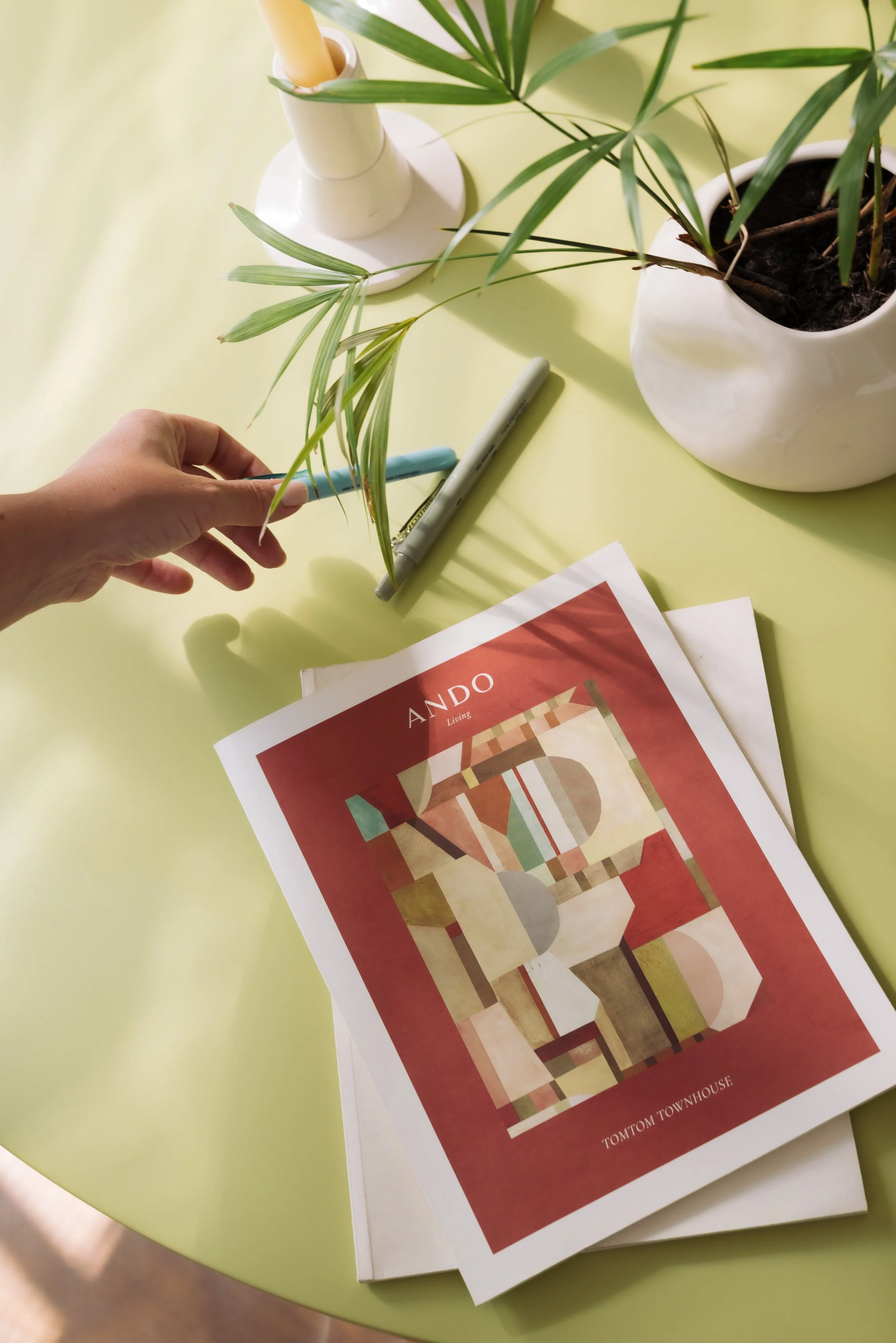 A hand holding a pen above a colorful art catalog on a light green table, with two potted plants nearby.