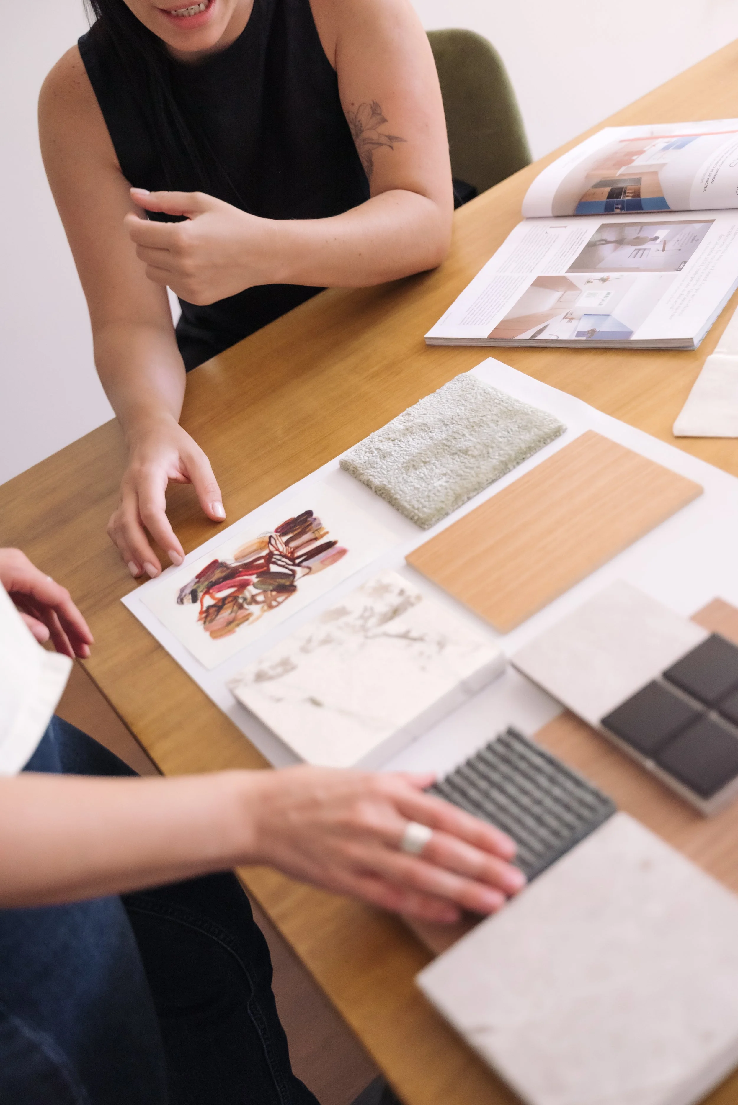 A person with a tattoo on their arm discussing interior design materials, including tiles and samples, at a wooden table.