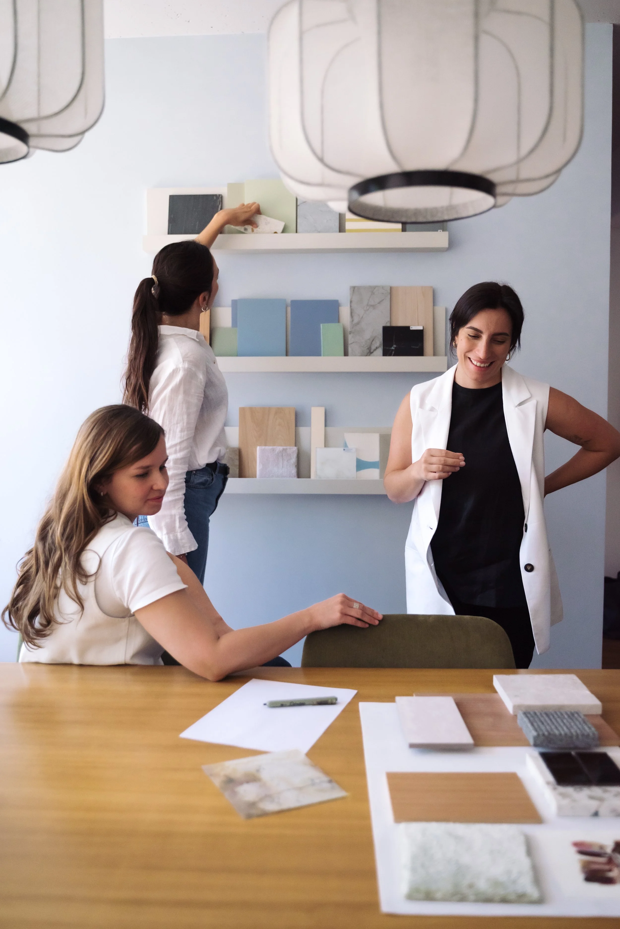 Three women in a design studio discussing tile samples, with one woman seated at a table and two women standing, one reaching for tile samples on a shelf.