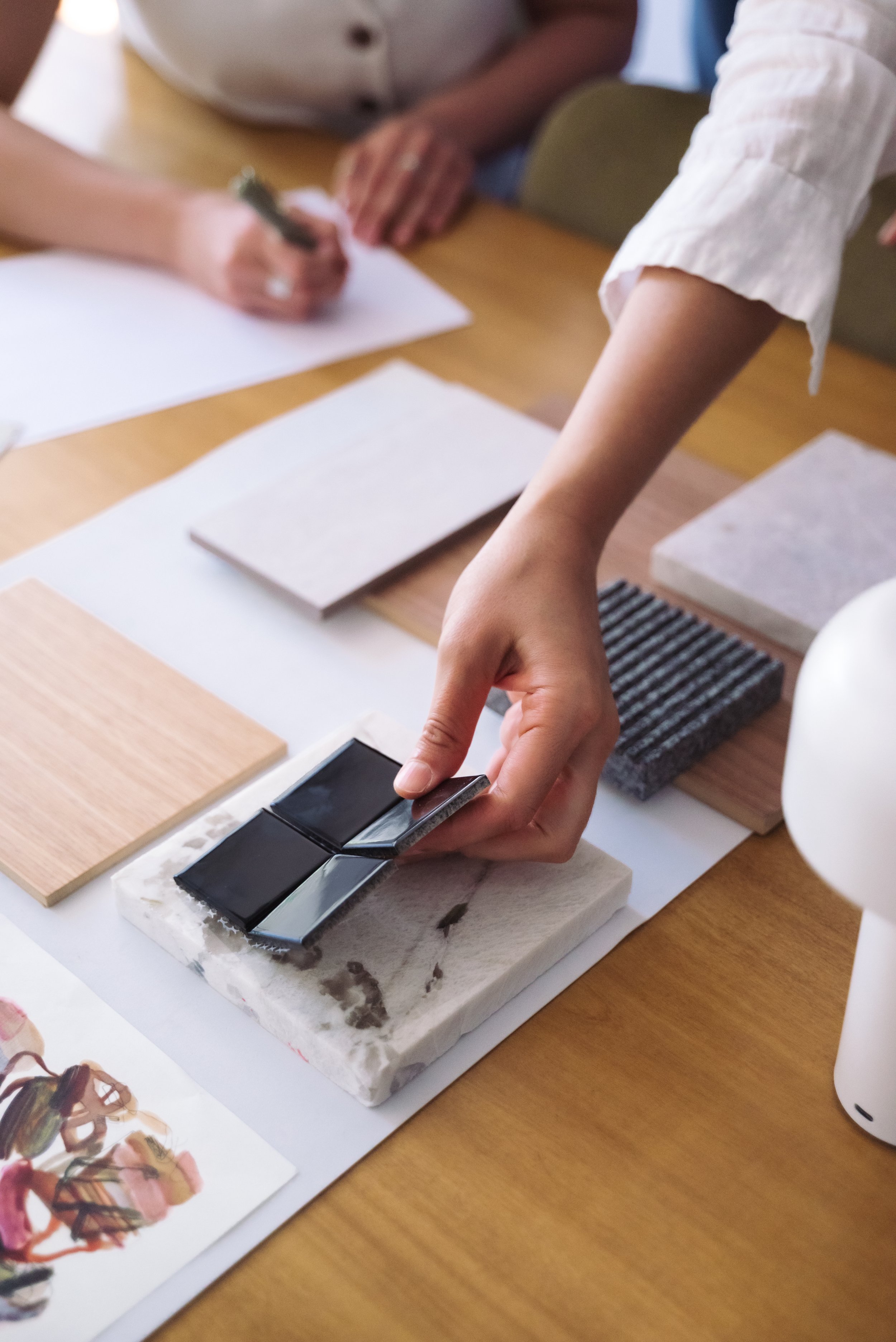 Person handling a color sample tile while other people work with paper and color swatches on a wooden table.