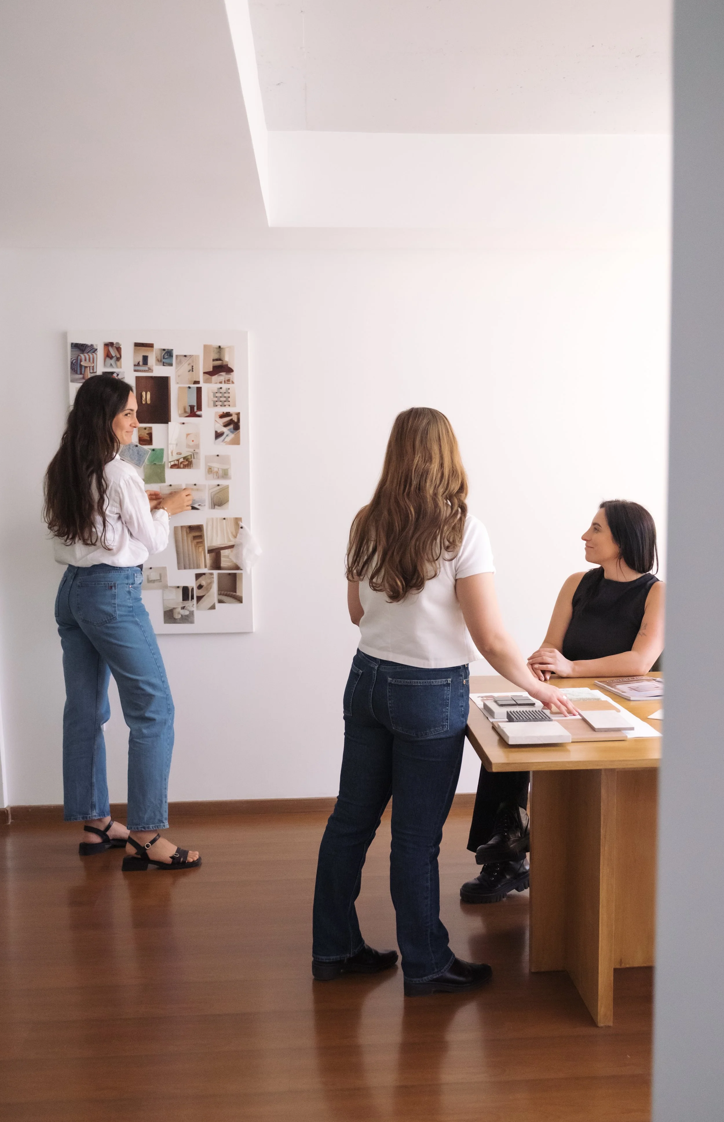 Three women discussing interior design options in a modern office with a white wall, a wooden desk, and mood boards.