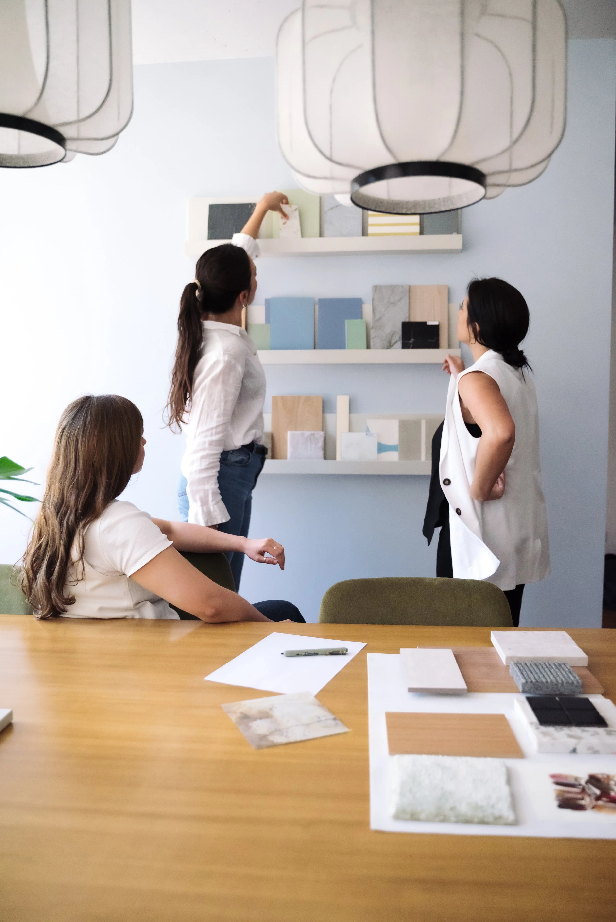 Three women in an interior design meeting discussing color and material samples on shelves and tables.