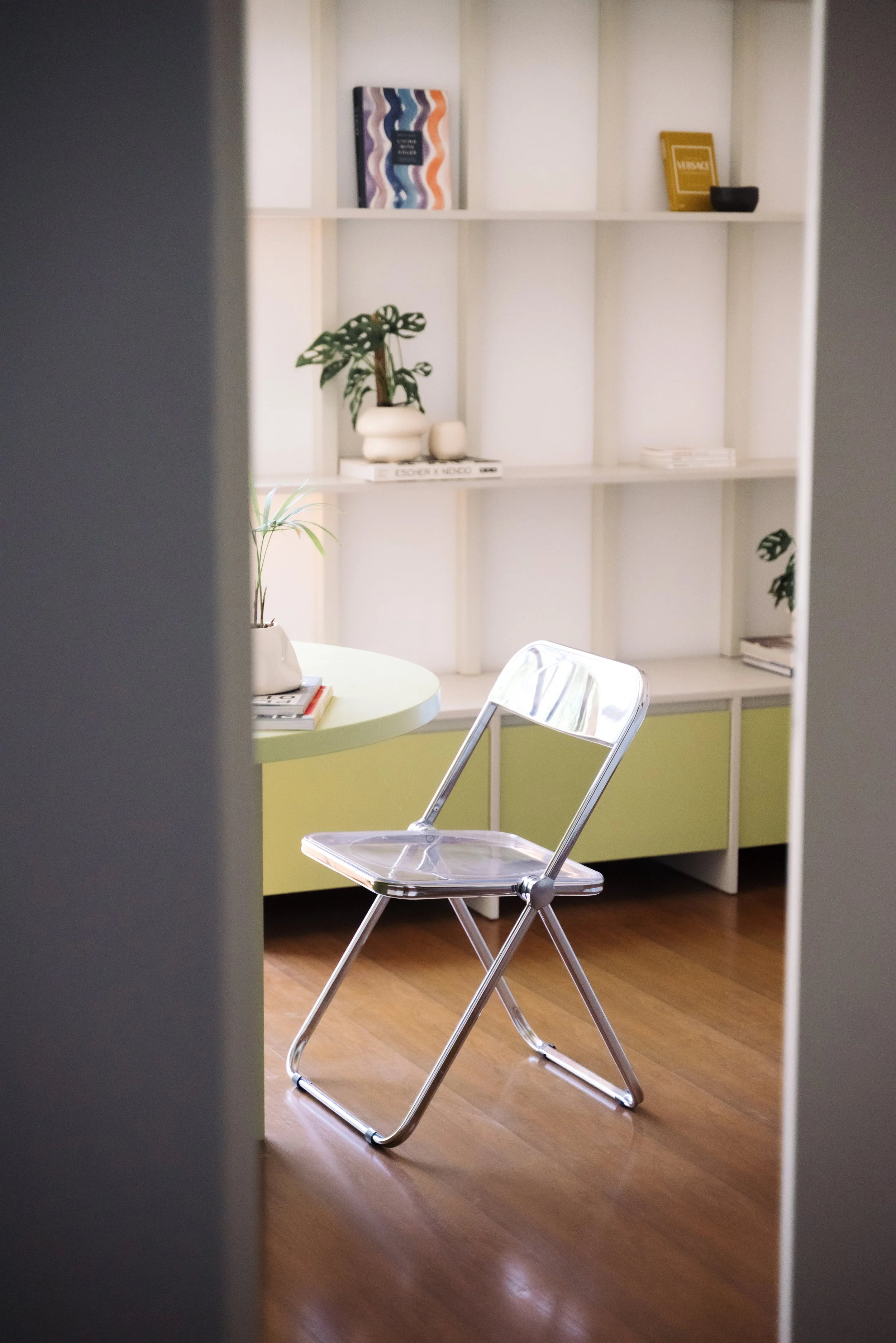 A modern office or sitting room with a white bookshelf, green cabinet, round table with books and plants, and a transparent folding chair on wooden floor, viewed through a doorway.