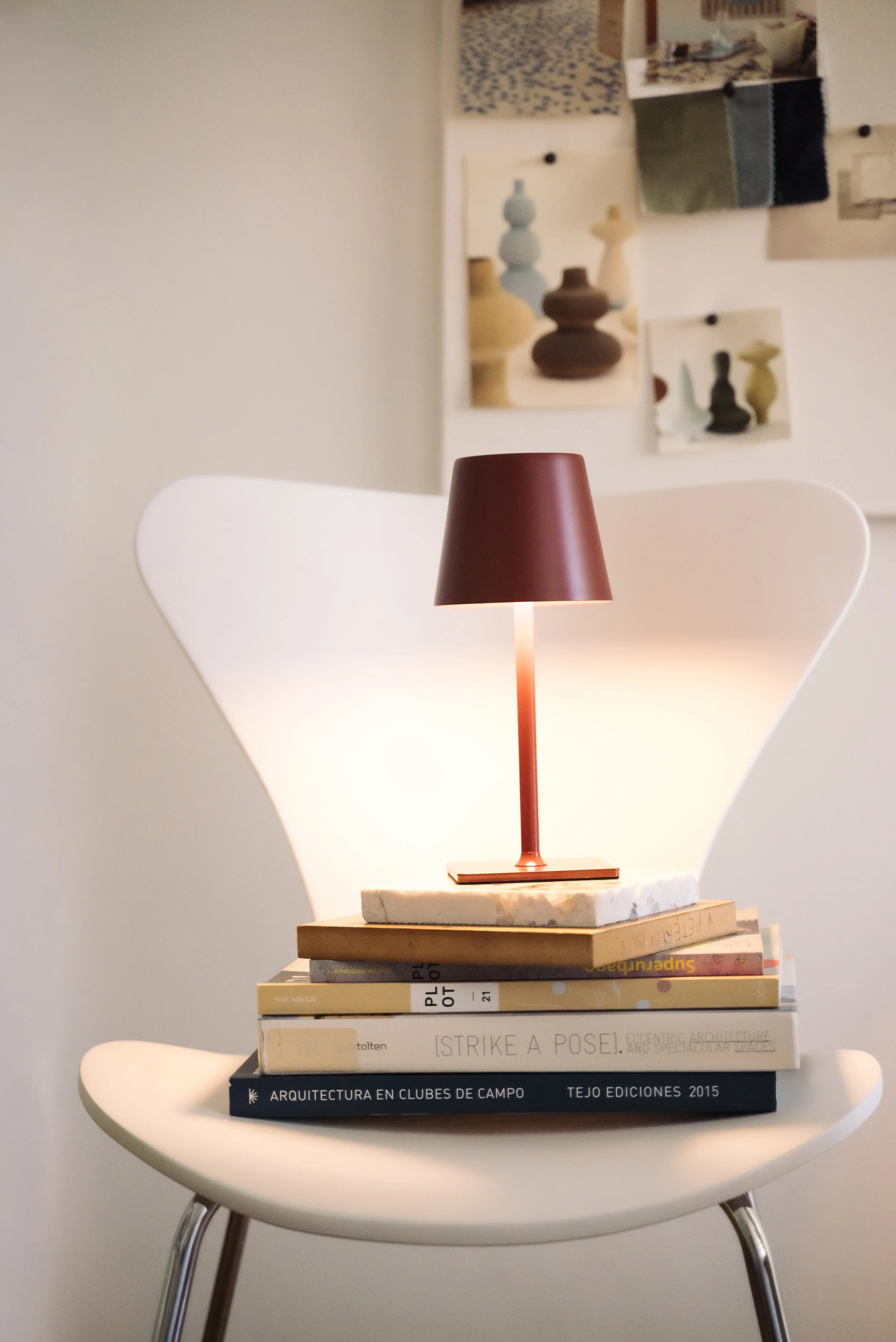 A modern scene featuring a white chair with a curved backrest, on which a stack of books is topped with a small, red desk lamp. The background shows a wall with several photographs and images pinned up.