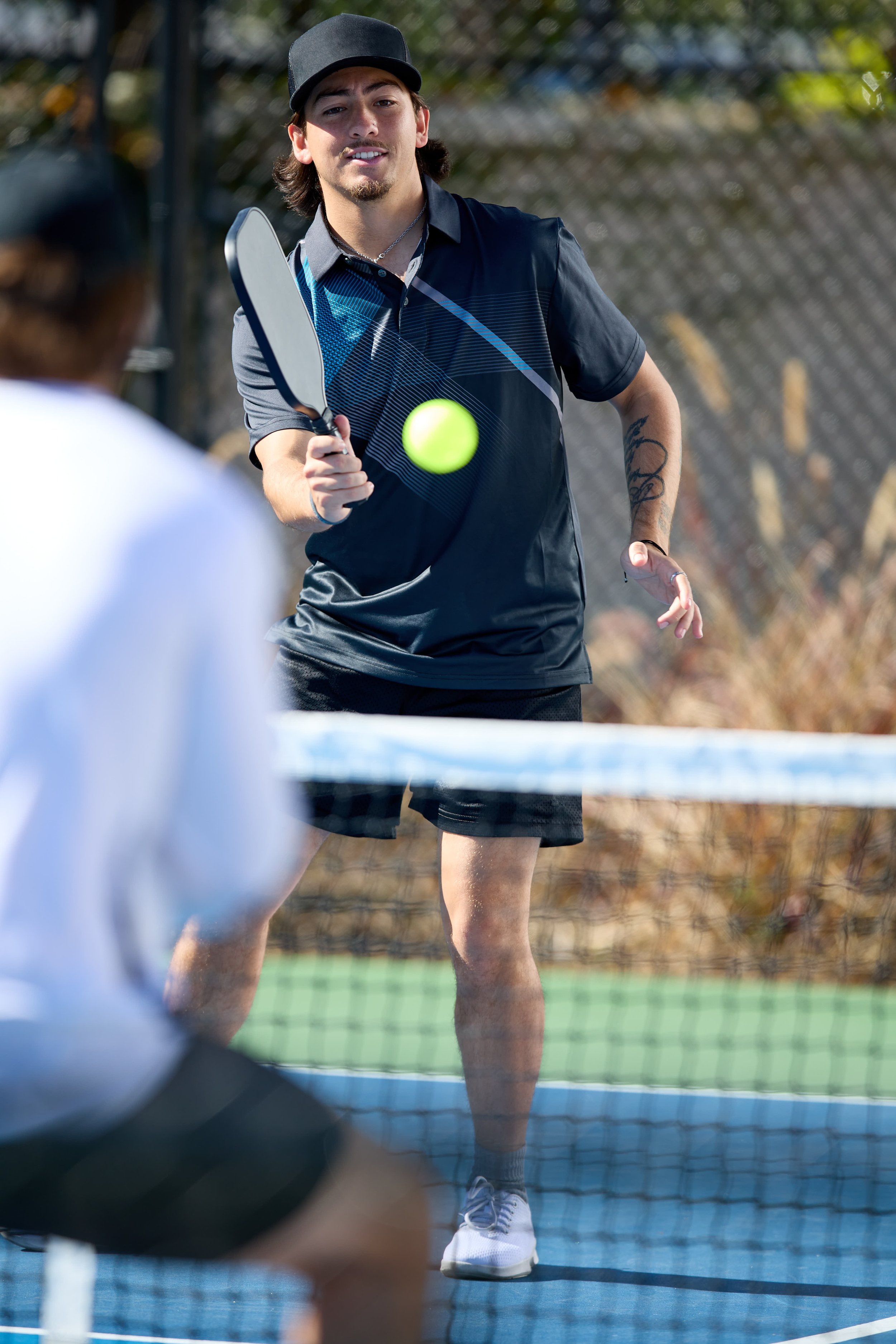 A young man wearing a black cap and sports outfit is playing pickleball, holding a paddle and ready to hit the yellow ball. An opponent is blurred in the foreground, and the net is visible.