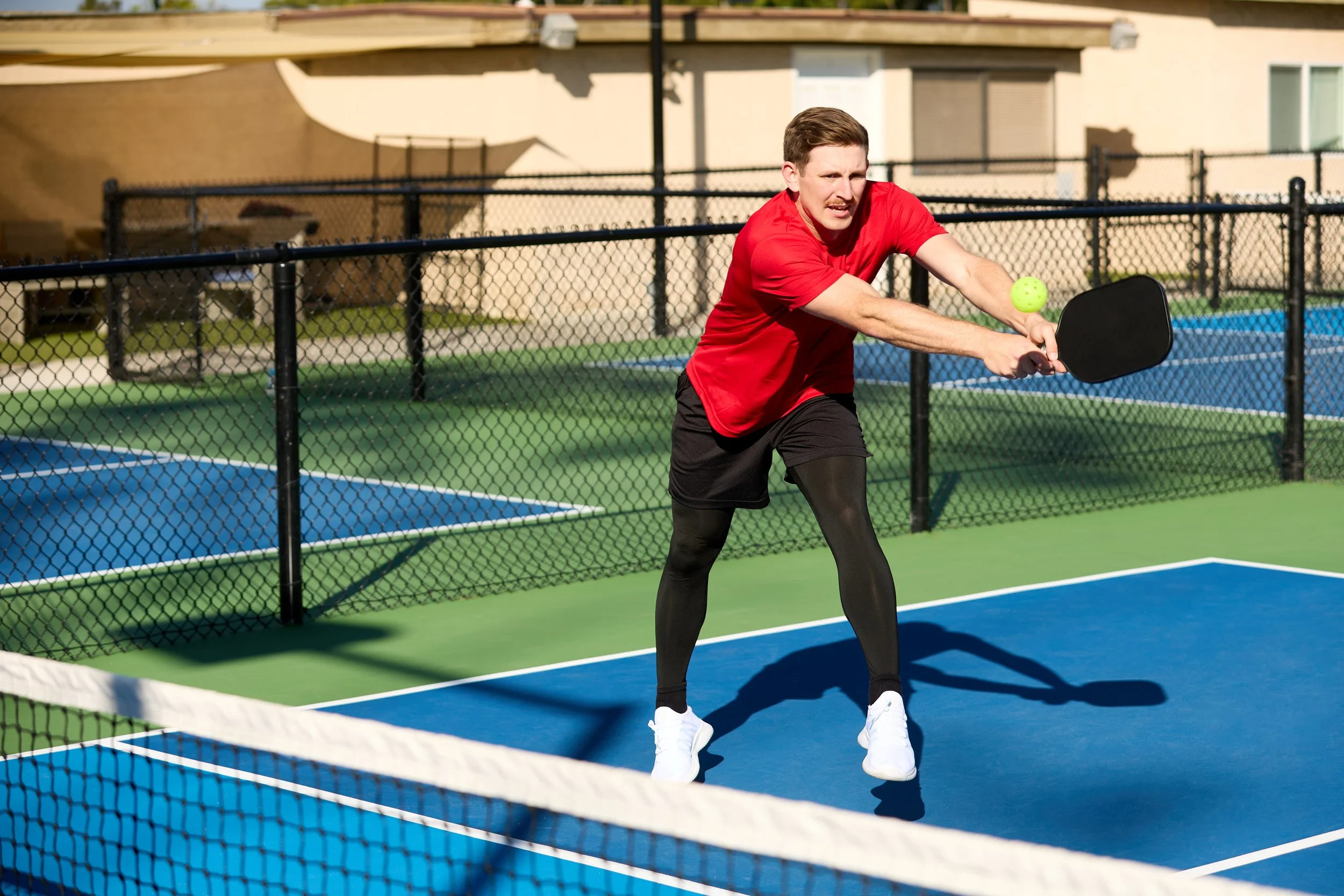 A man playing pickleball on a court with a green and blue surface, hitting a green ball with a black paddle, wearing a red shirt, black shorts, black leggings, and white shoes.