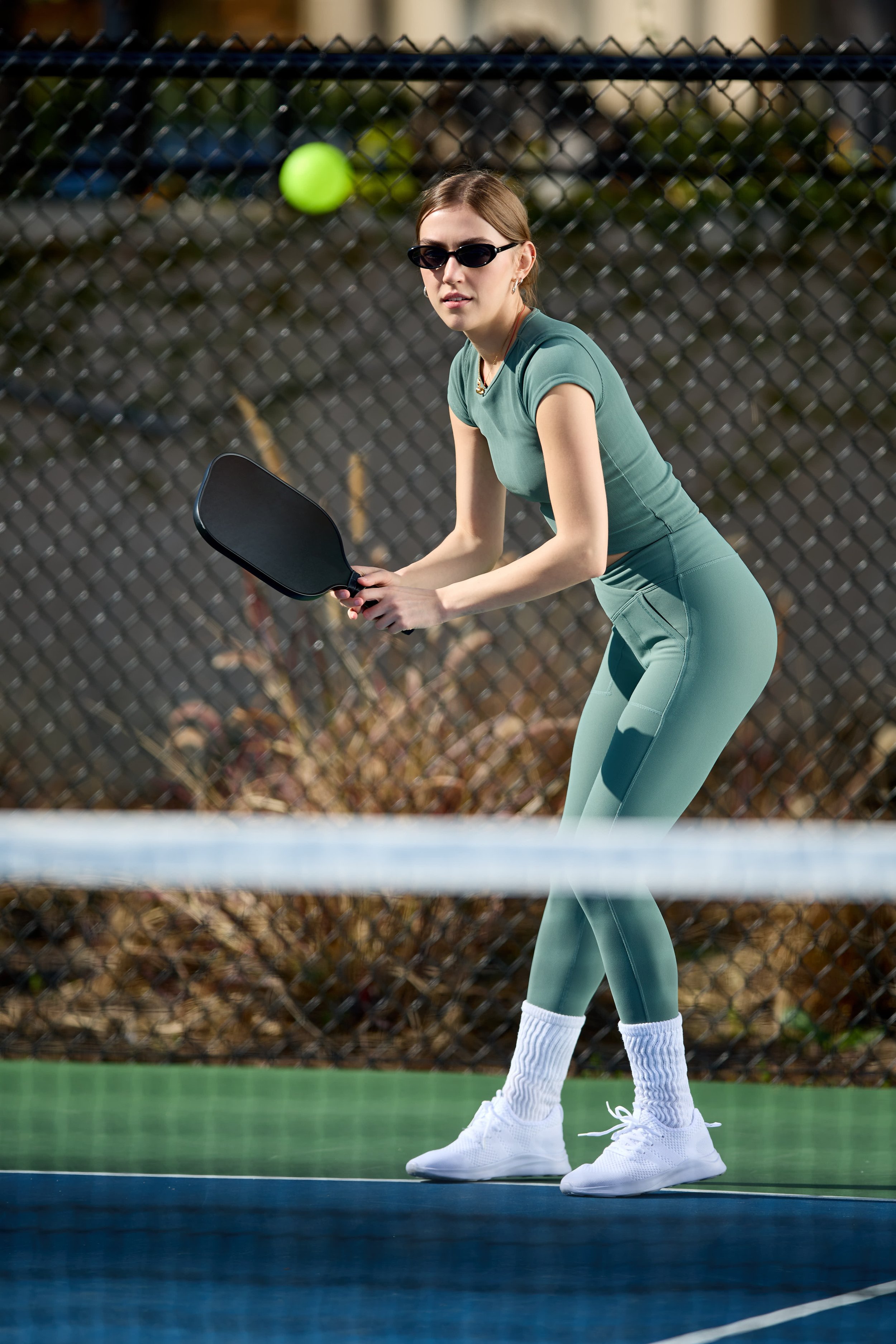 A woman wearing sunglasses, a teal athletic outfit, white socks, and white sneakers playing pickleball on an outdoor court with a chain-link fence in the background, preparing to hit a tennis-like ball with a paddle.