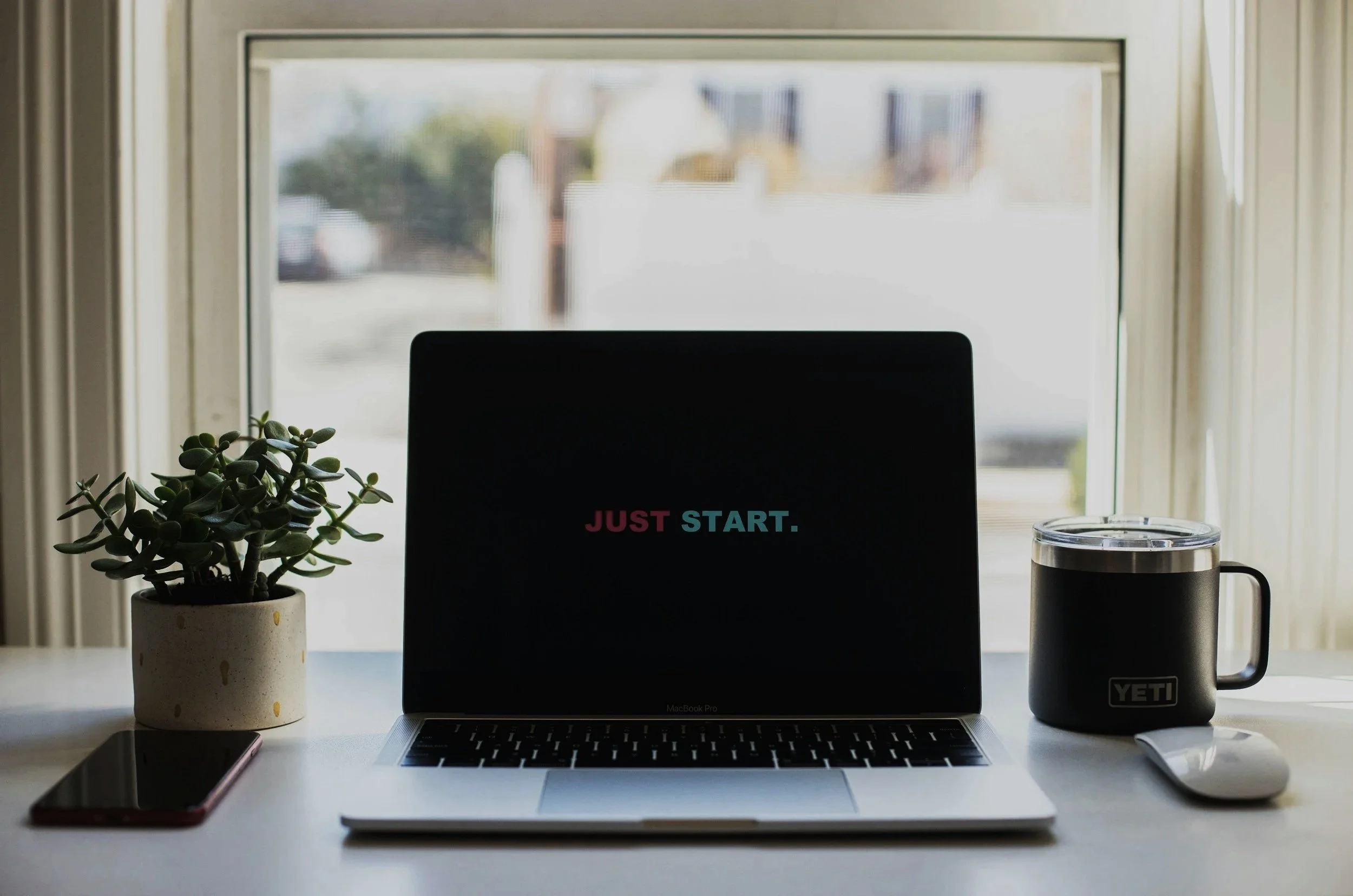 A workspace with a MacBook Pro laptop displaying the words 'JUST START' on the screen, a potted succulent plant, an iPhone, a Yeti mug, and a reusable water bottle on a white desk in front of a window with beige curtains.