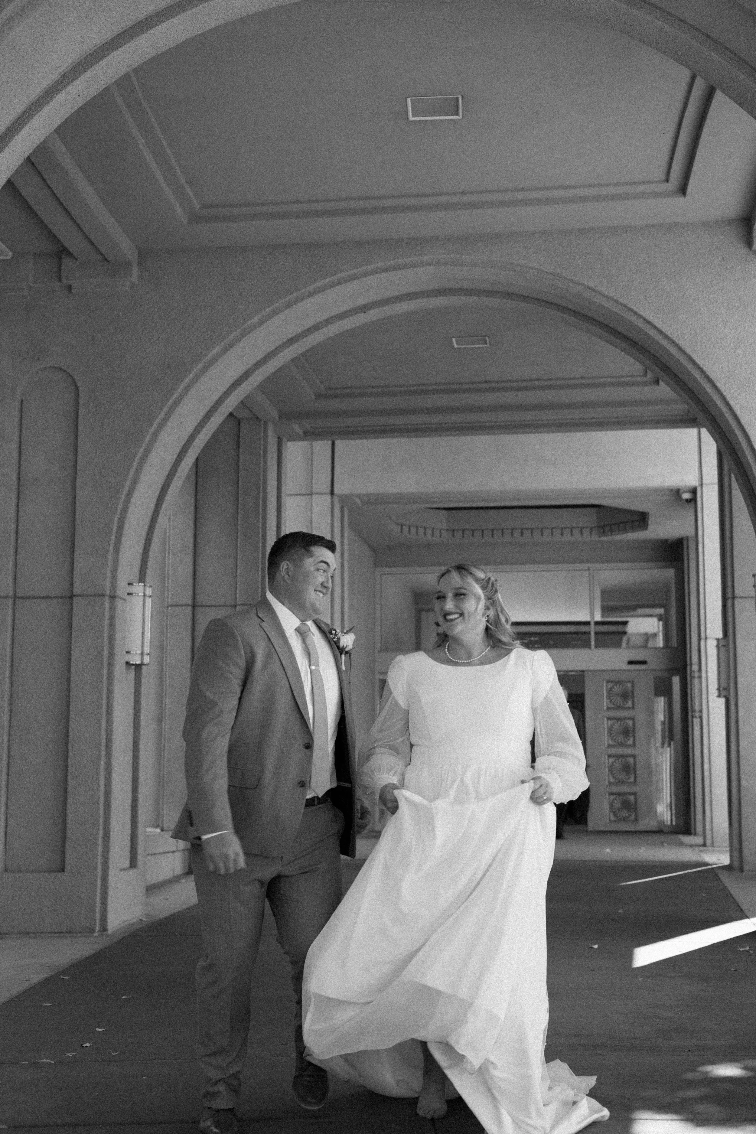 A black and white photo of a smiling couple in wedding attire walking together under a decorative archway, with the bride holding up her wedding dress.