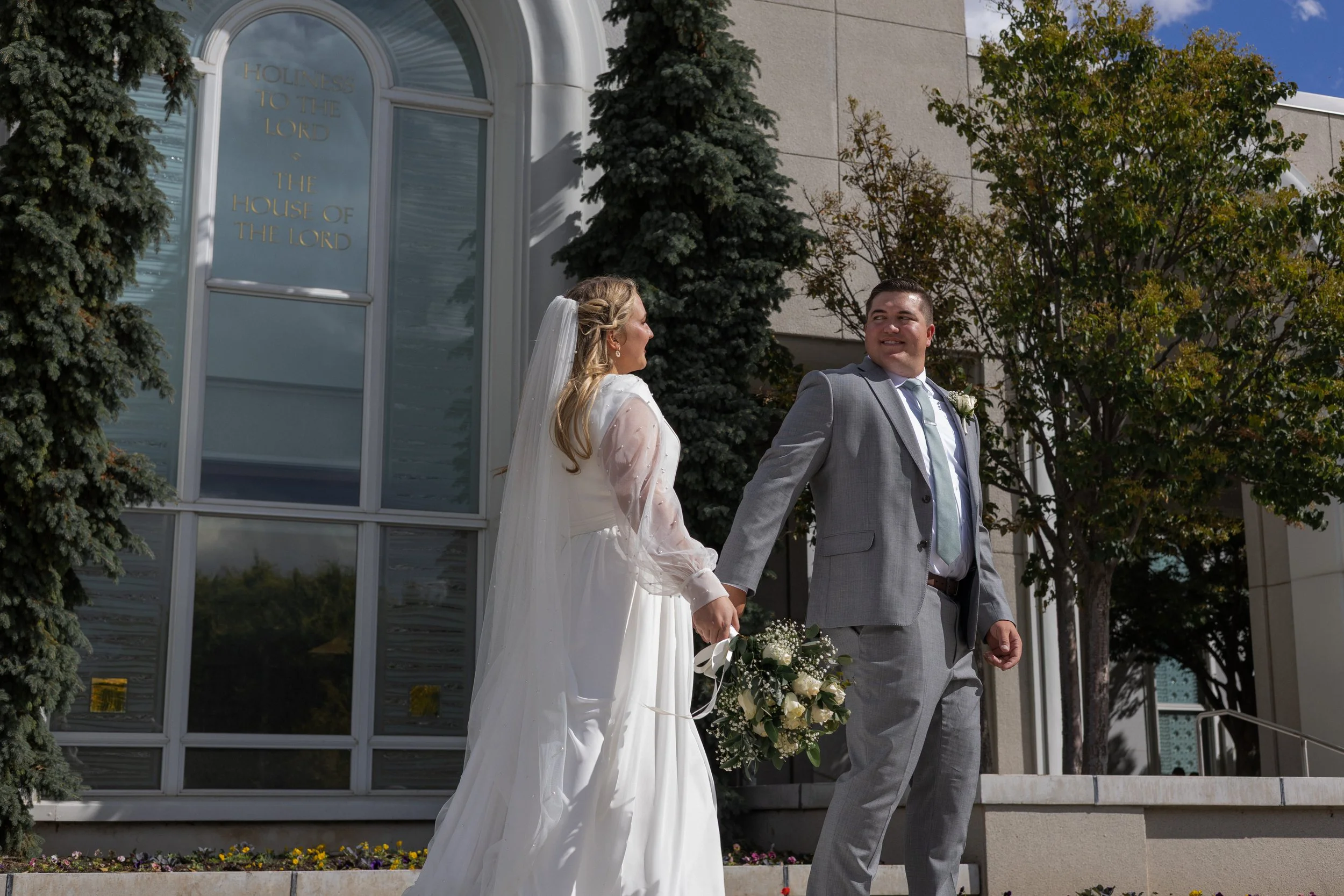 A bride and groom holding hands outside a church, with the bride dressed in a white gown and the groom in a gray suit, smiling at each other.