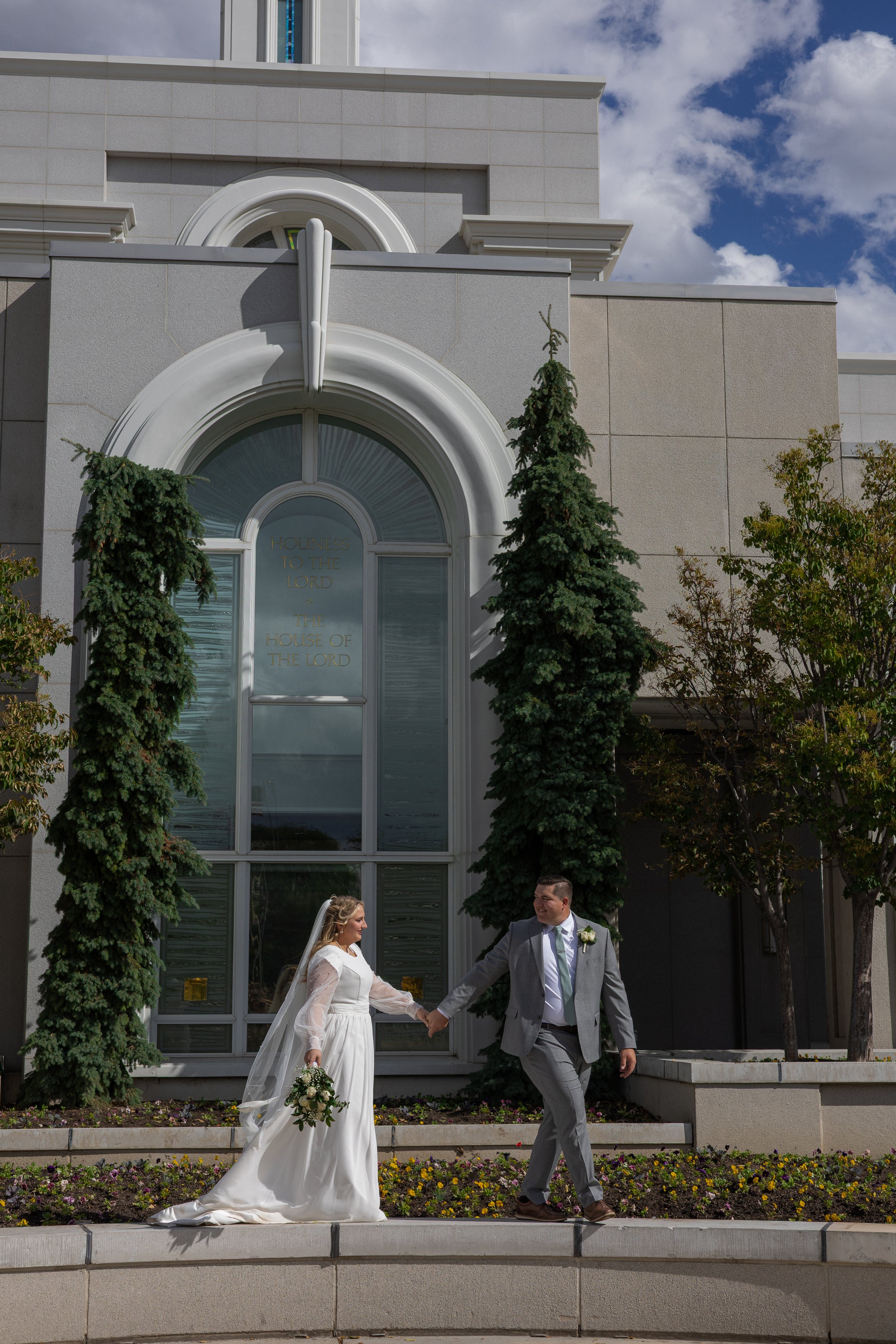 A bride and groom holding hands outside a church with a large arched window and tall evergreen trees.