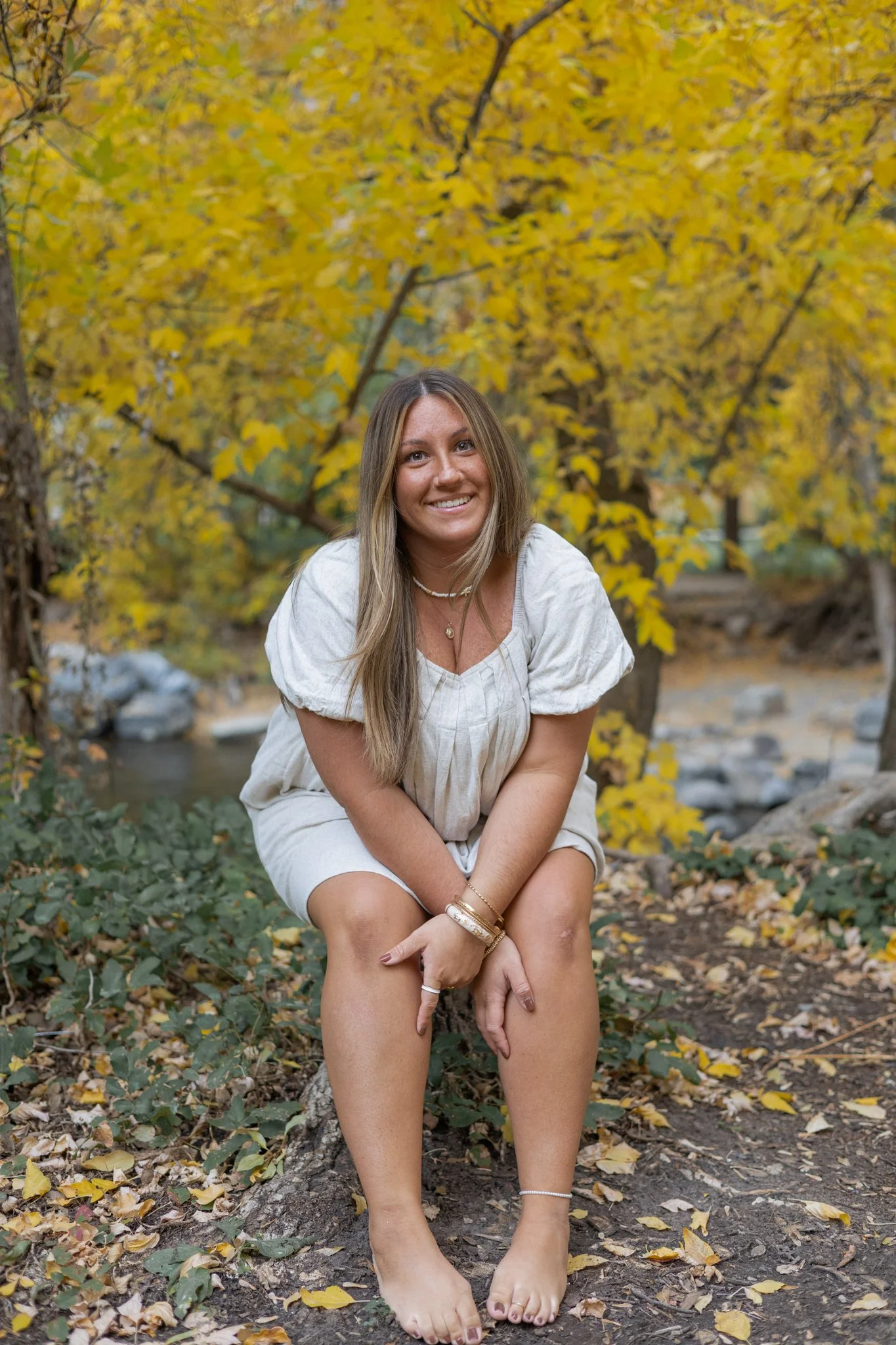 A woman with long blonde hair is sitting on a tree stump in a forest with yellow fall foliage, smiling at the camera.