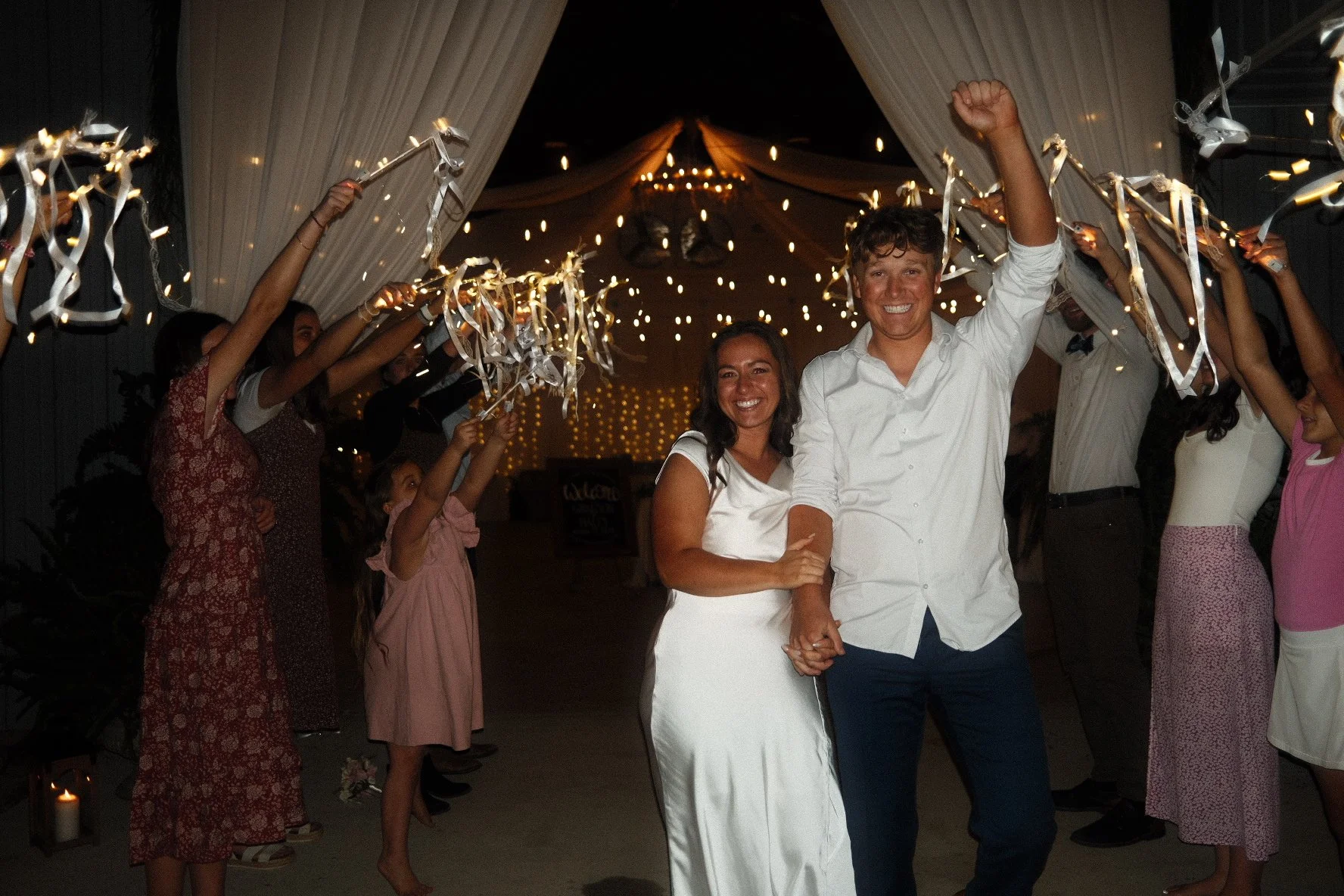 A newlywed couple in wedding attire celebrating with friends and family at night under a decorated tent with string lights, people holding ribbons in a celebratory procession.