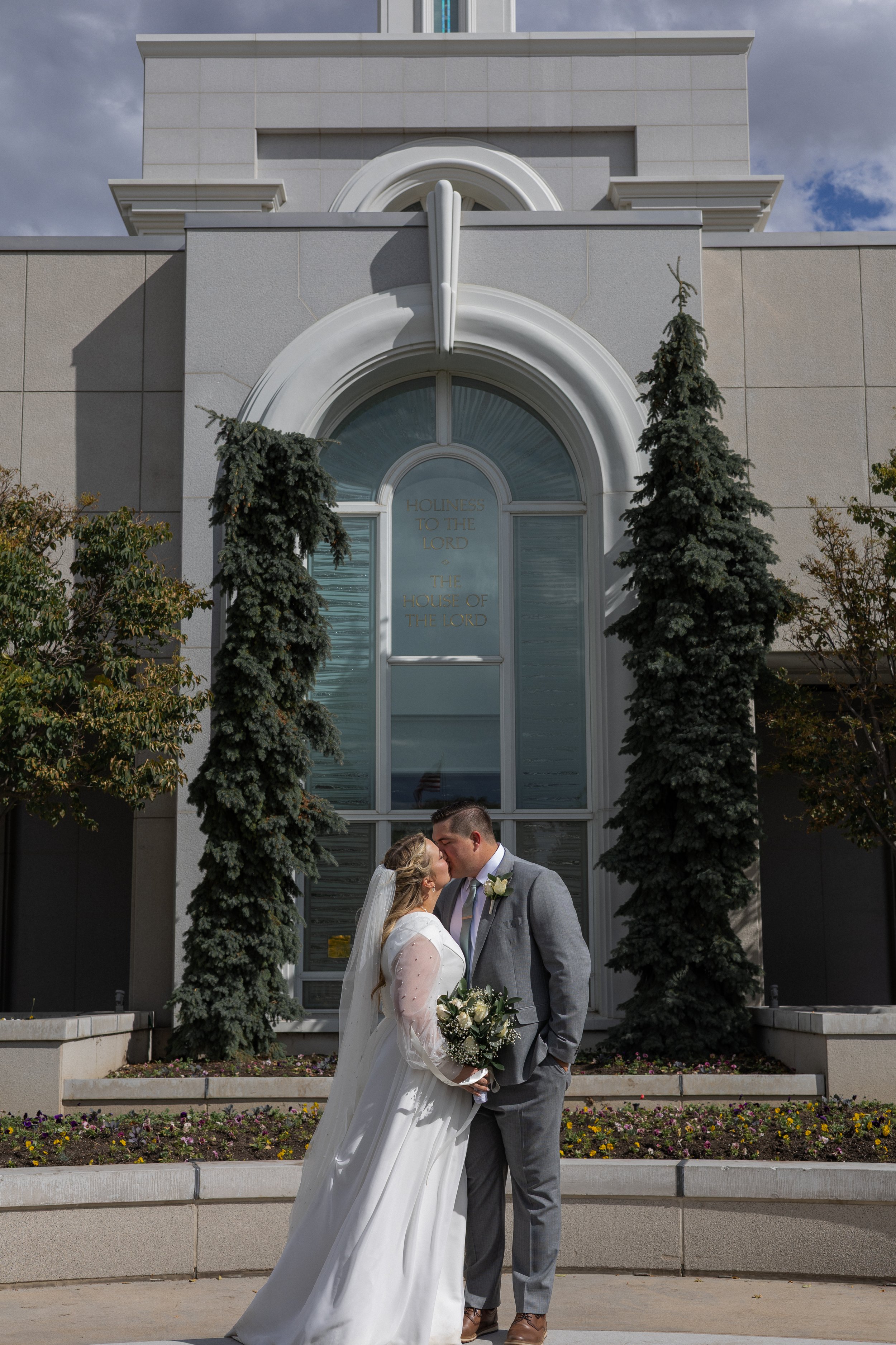 A bride and groom sharing a kiss in front of a church building with large arched windows and tall evergreen trees.