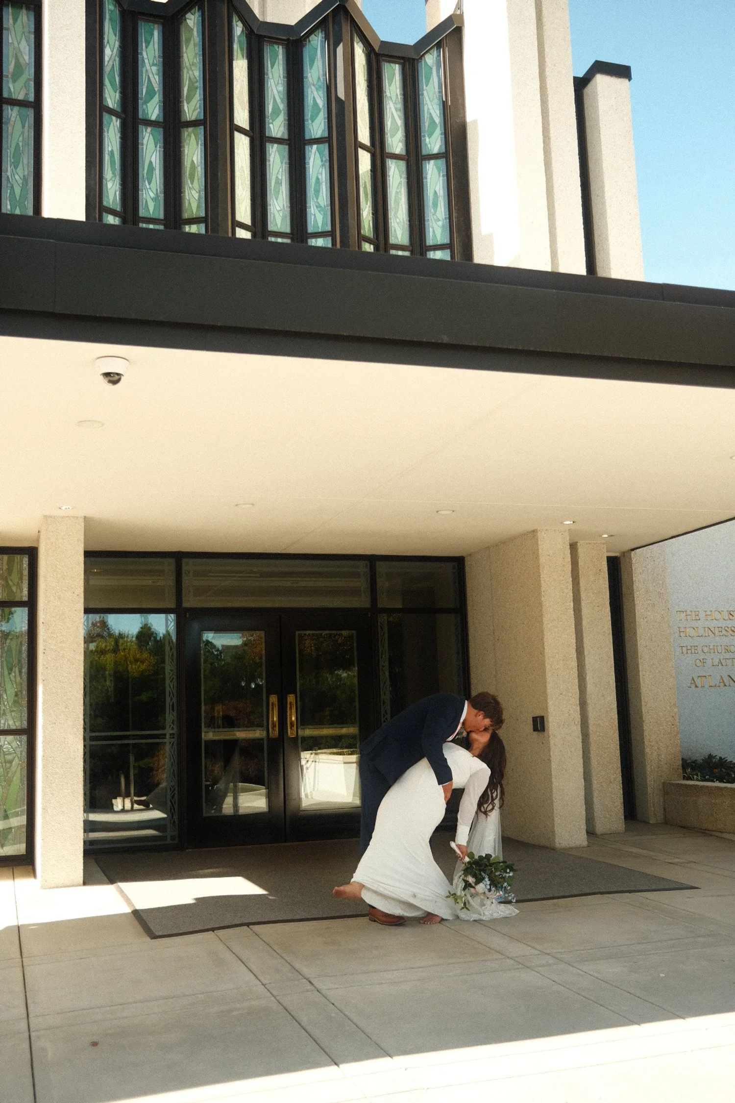 A bride and groom sharing a kiss outside a building, with the groom lifting the bride's veil, holding a bouquet of flowers.