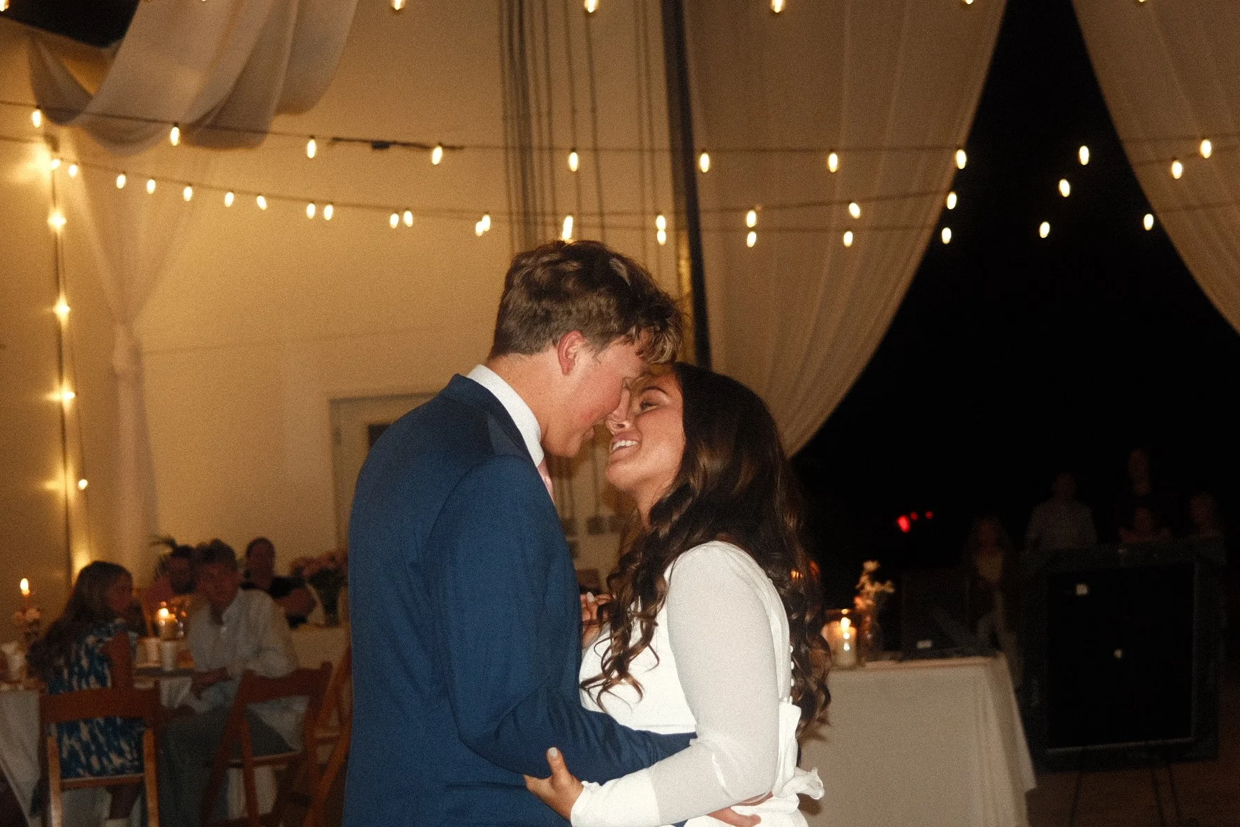 A couple dancing at a wedding reception under string lights, smiling and looking into each other's eyes.