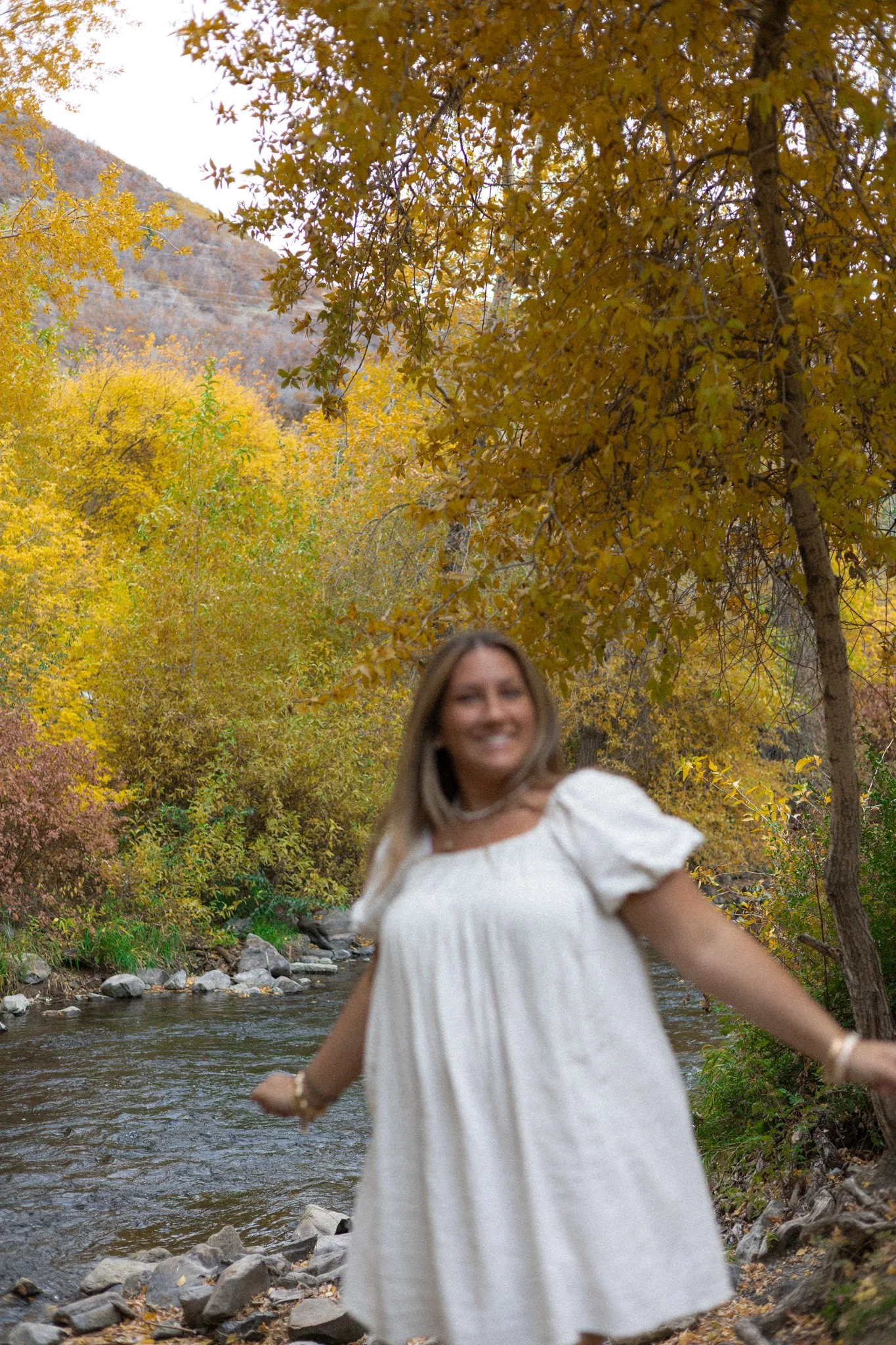 A woman in a white dress standing by a river surrounded by colorful autumn trees with yellow and orange leaves.