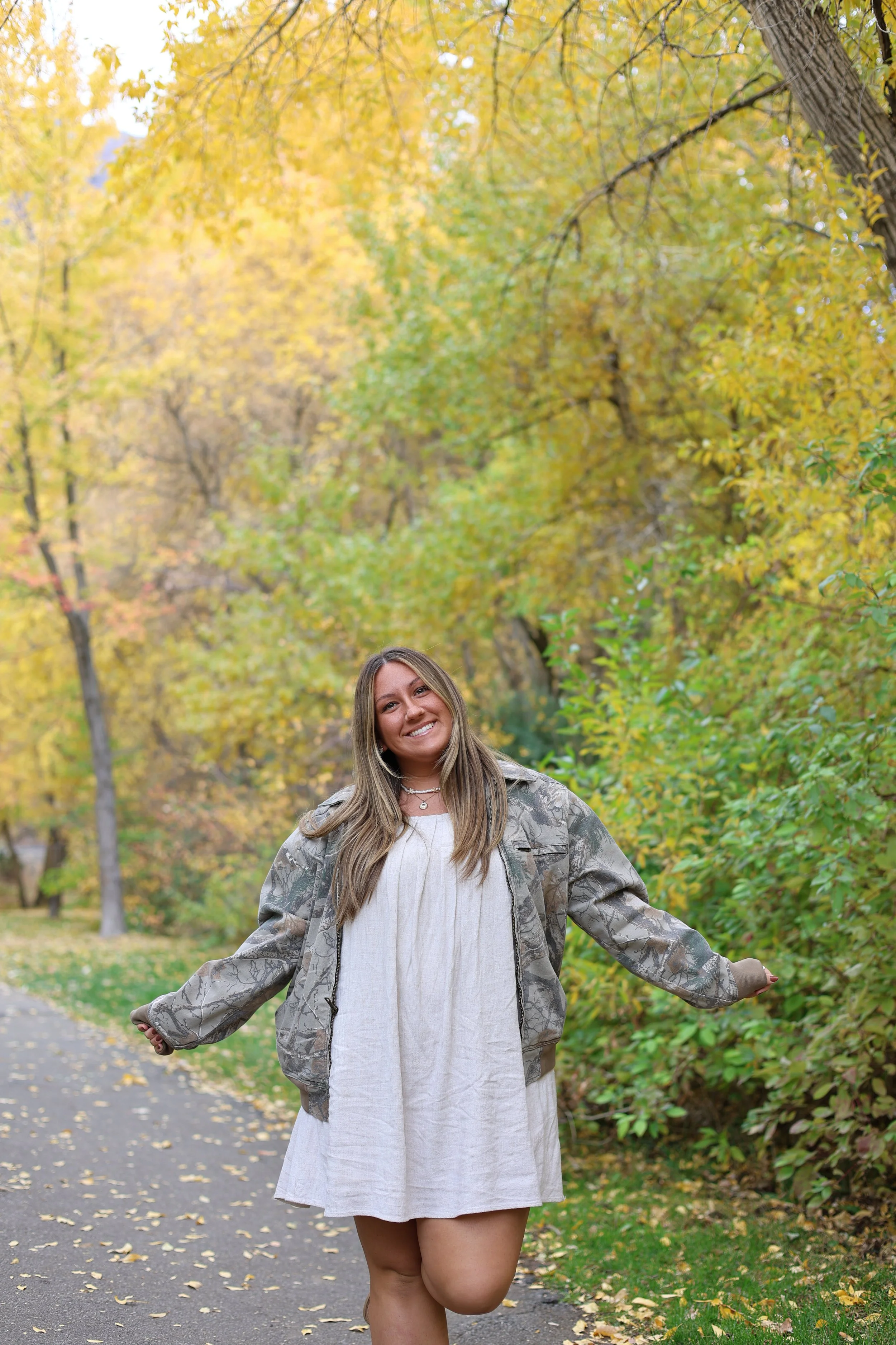 A smiling young woman walking on a path in a park during fall, surrounded by yellow and orange autumn leaves.