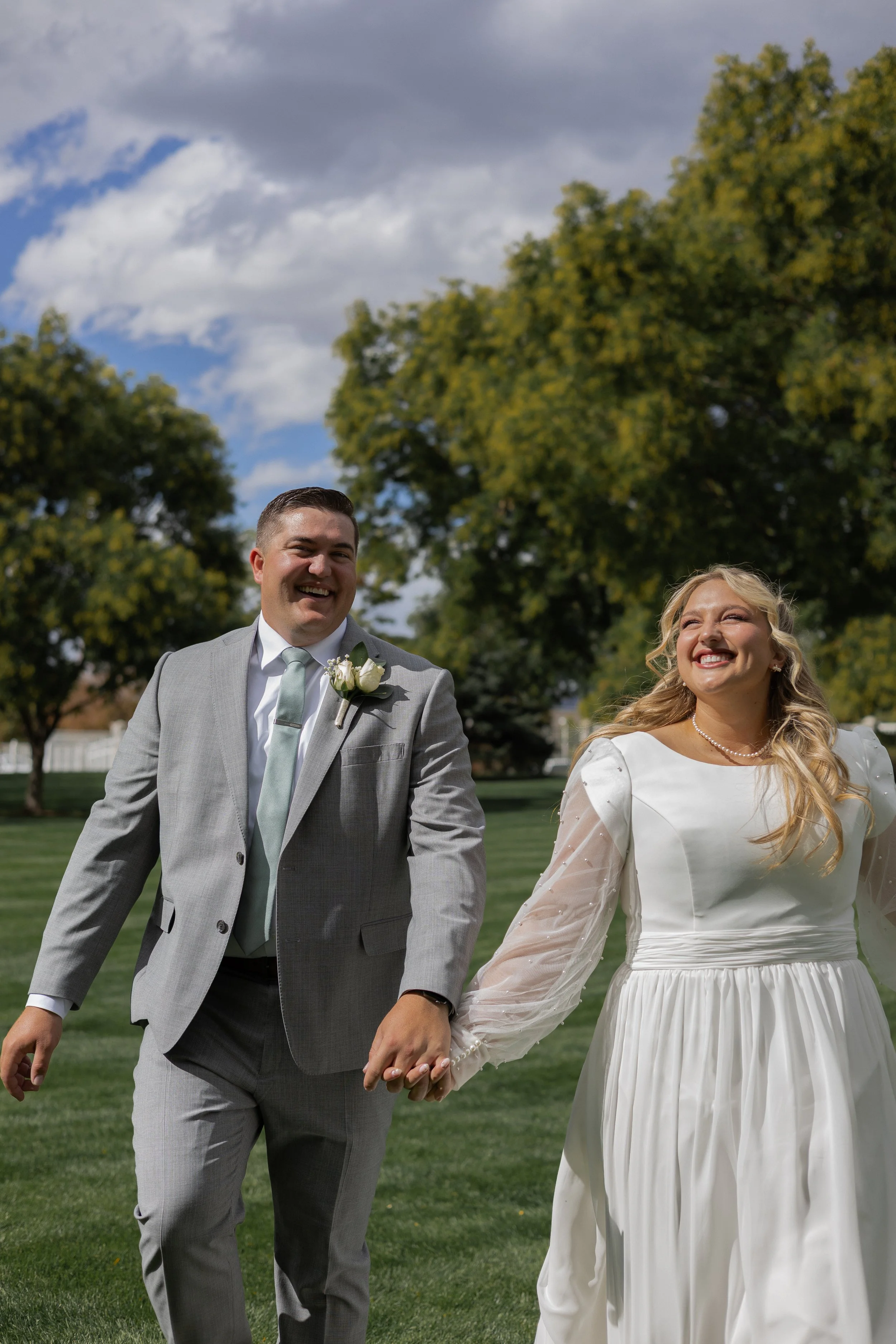 A smiling bride and groom holding hands outdoors on a sunny day with scattered clouds, trees, and a lush green lawn in the background.