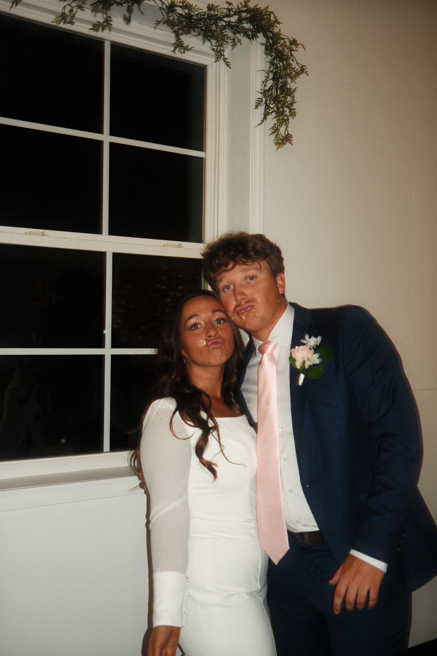 A woman and a man making silly faces in a dimly lit room at night, standing in front of a white wall and window, dressed in formal attire.