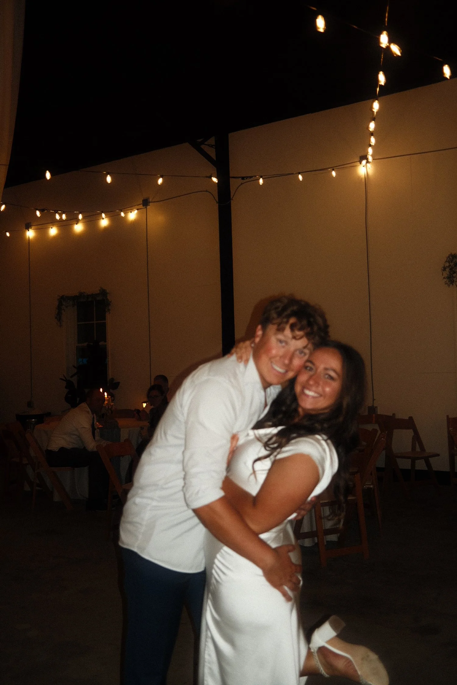 A couple smiling and hugging at an evening event with string lights hanging above them in an outdoor setting.