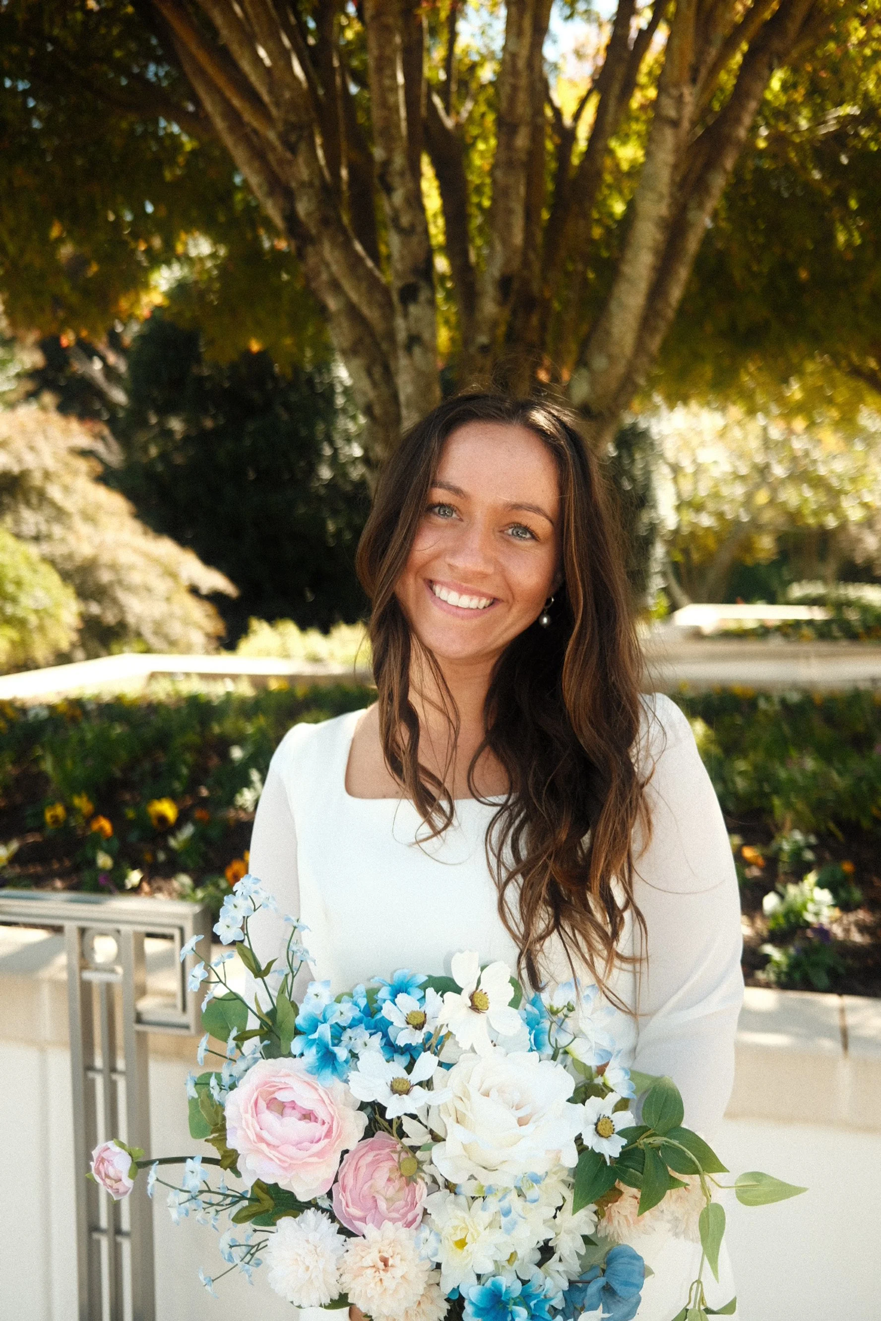 A woman with long wavy brown hair and blue eyes, smiling while holding a bouquet of flowers outdoors under a large tree with green and yellow leaves.