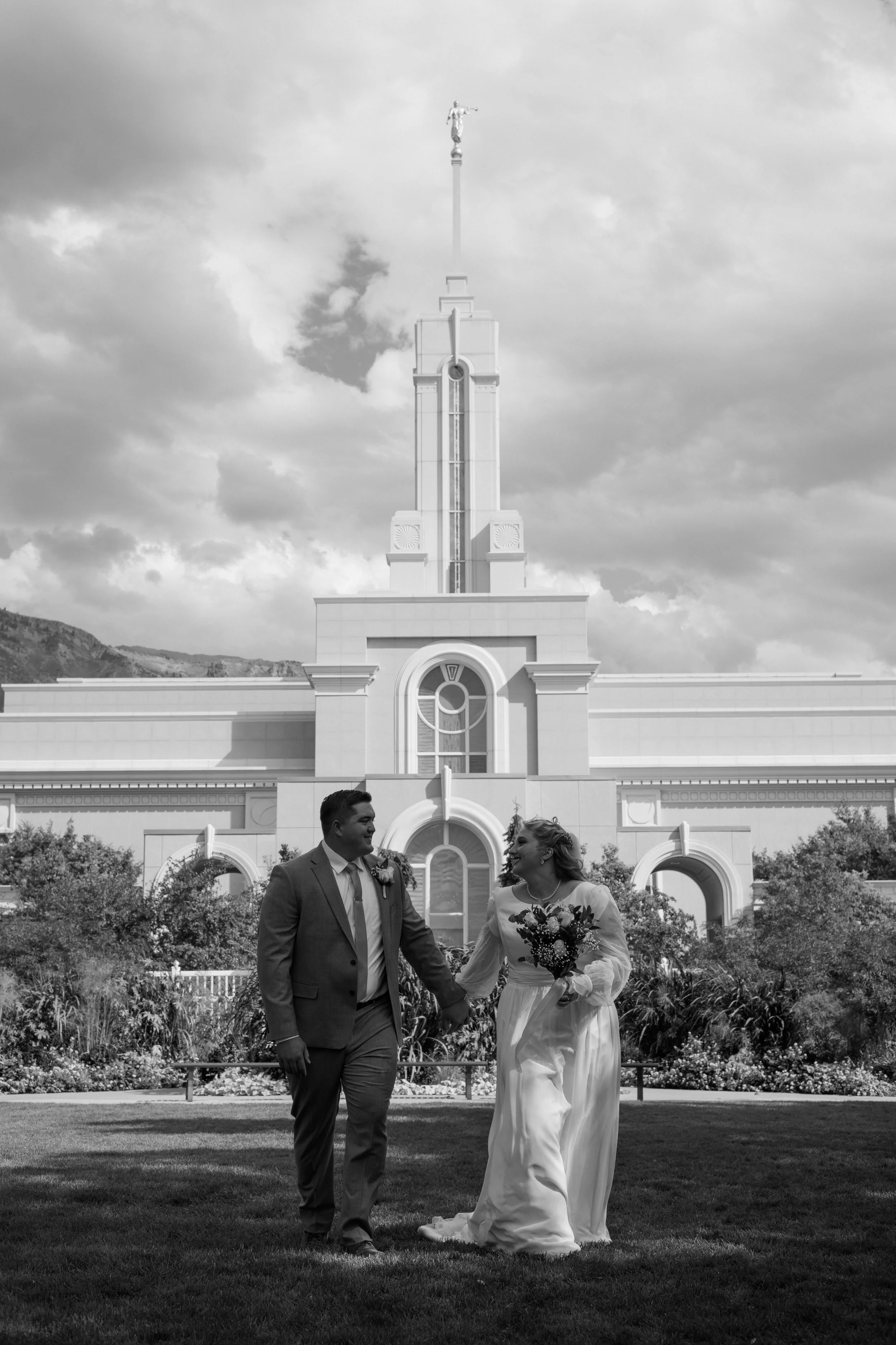 A black and white photo of a smiling couple holding hands and walking on a grassy area in front of a large, ornate building with a tall tower topped by a statue. The woman is wearing a long dress and holding a bouquet, while the man is dressed in a s