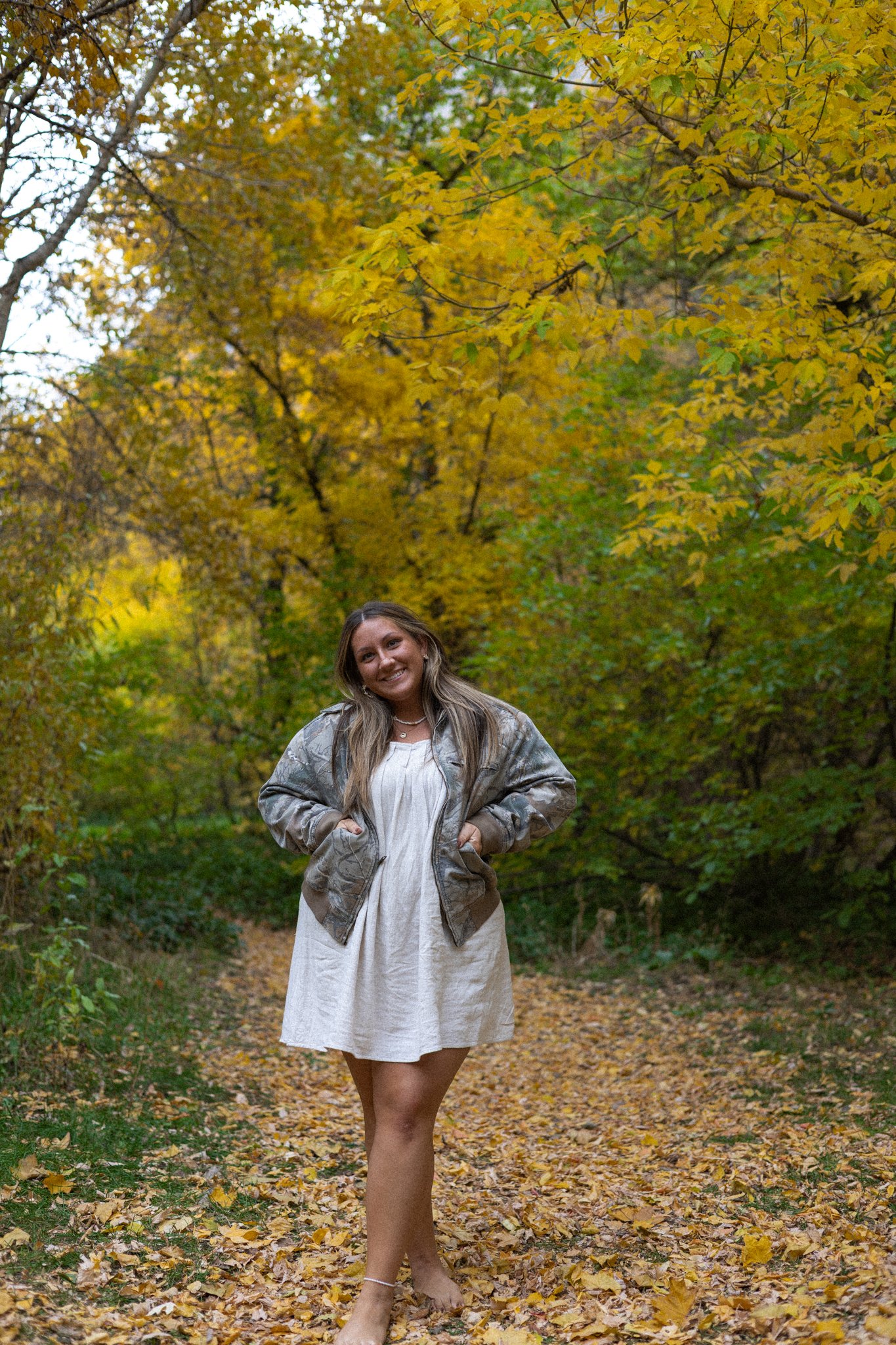 A woman standing on a leaf-covered trail in a forest with autumn foliage, smiling at the camera, wearing a beige dress, a camouflage jacket, and barefoot.
