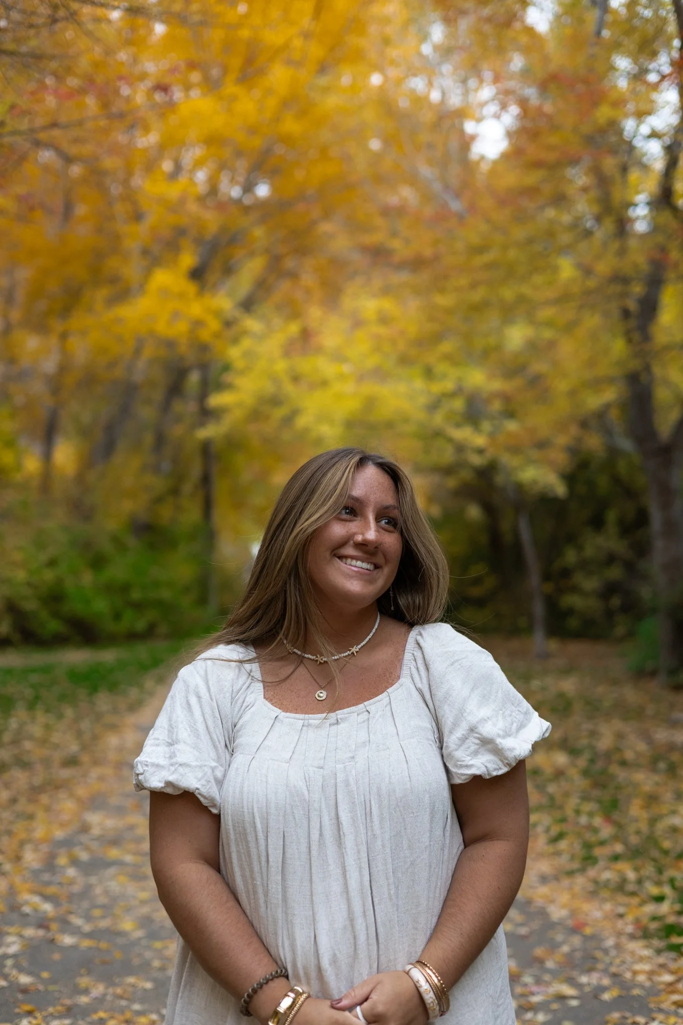 A woman smiling outdoors surrounded by yellow and orange autumn trees, wearing a white dress with jewelry.