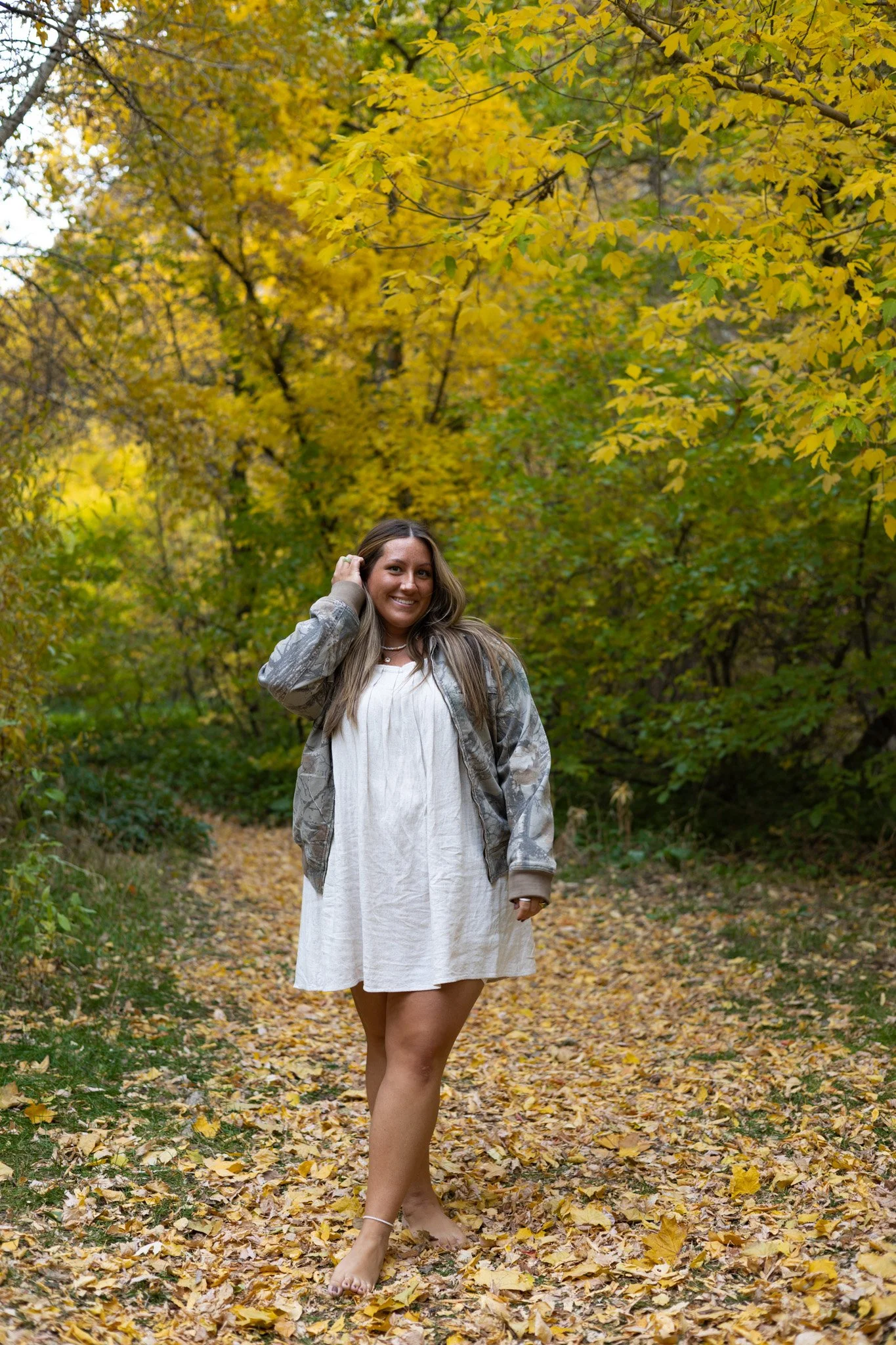 A woman smiling and standing barefoot on a leaf-covered trail in a forest with yellow autumn foliage.