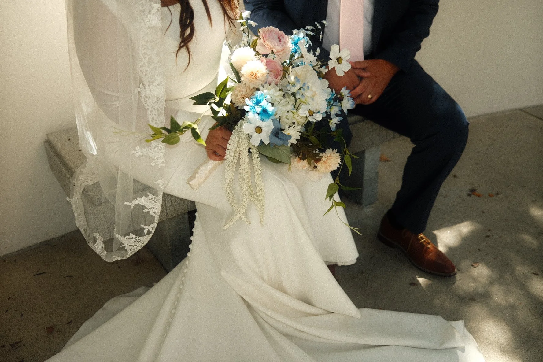 Close-up of a bride holding a bouquet of pink, white, blue, and green flowers, sitting on a bench next to a groom dressed in a dark suit. The bride's white dress has lace details and buttons on the back.
