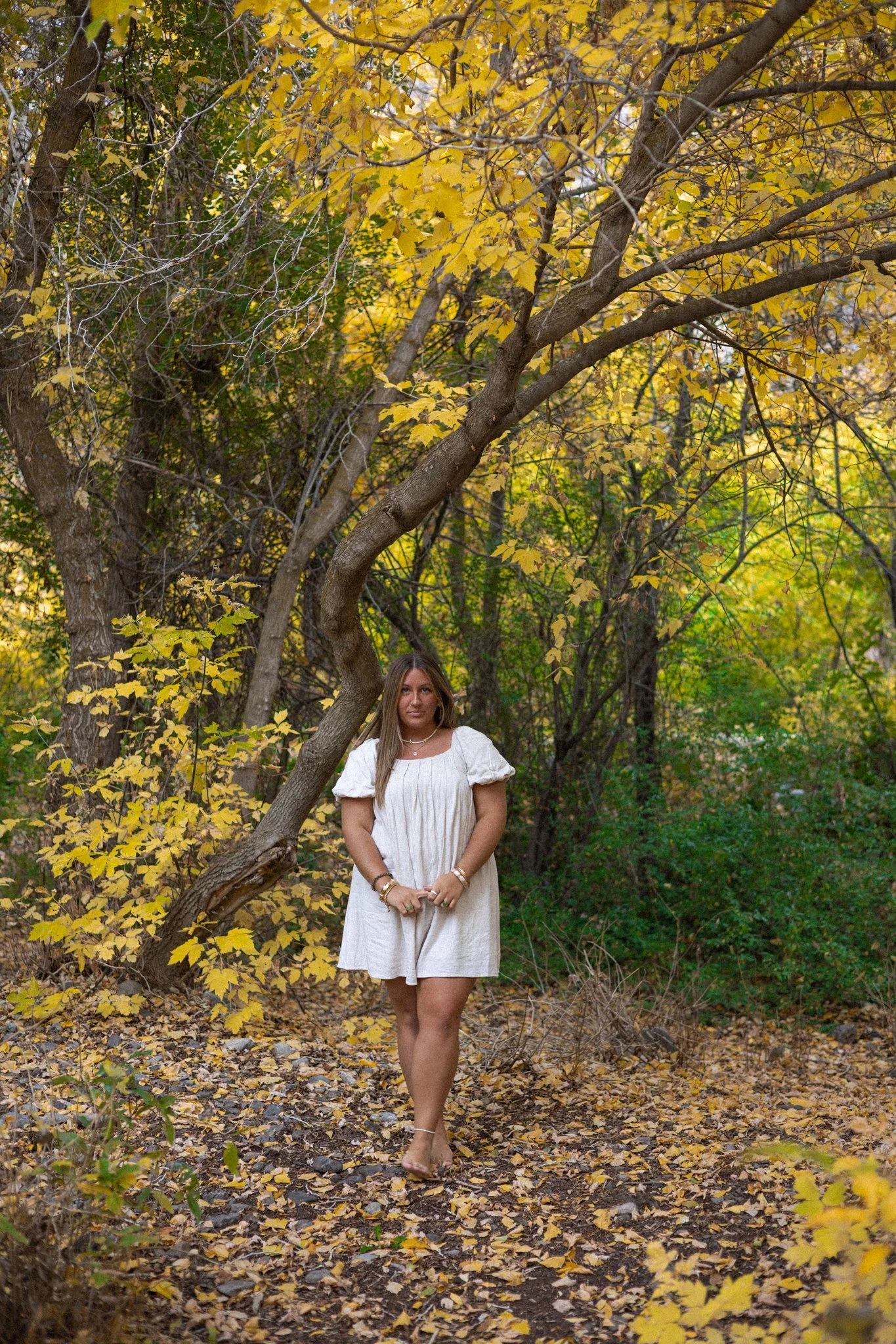 Woman in a white dress standing on a leaf-covered trail in a wooded area with yellow and green autumn foliage.