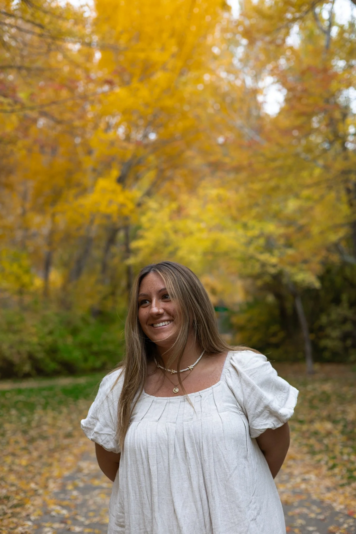 A woman with long, light brown hair smiling outdoors in autumn, standing on a path surrounded by fall foliage with trees showing yellow and orange leaves.