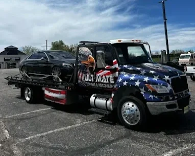 A flatbed tow truck decorated with an American flag theme, carrying a black sedan in its bed, parked in a lot under a partly cloudy sky.