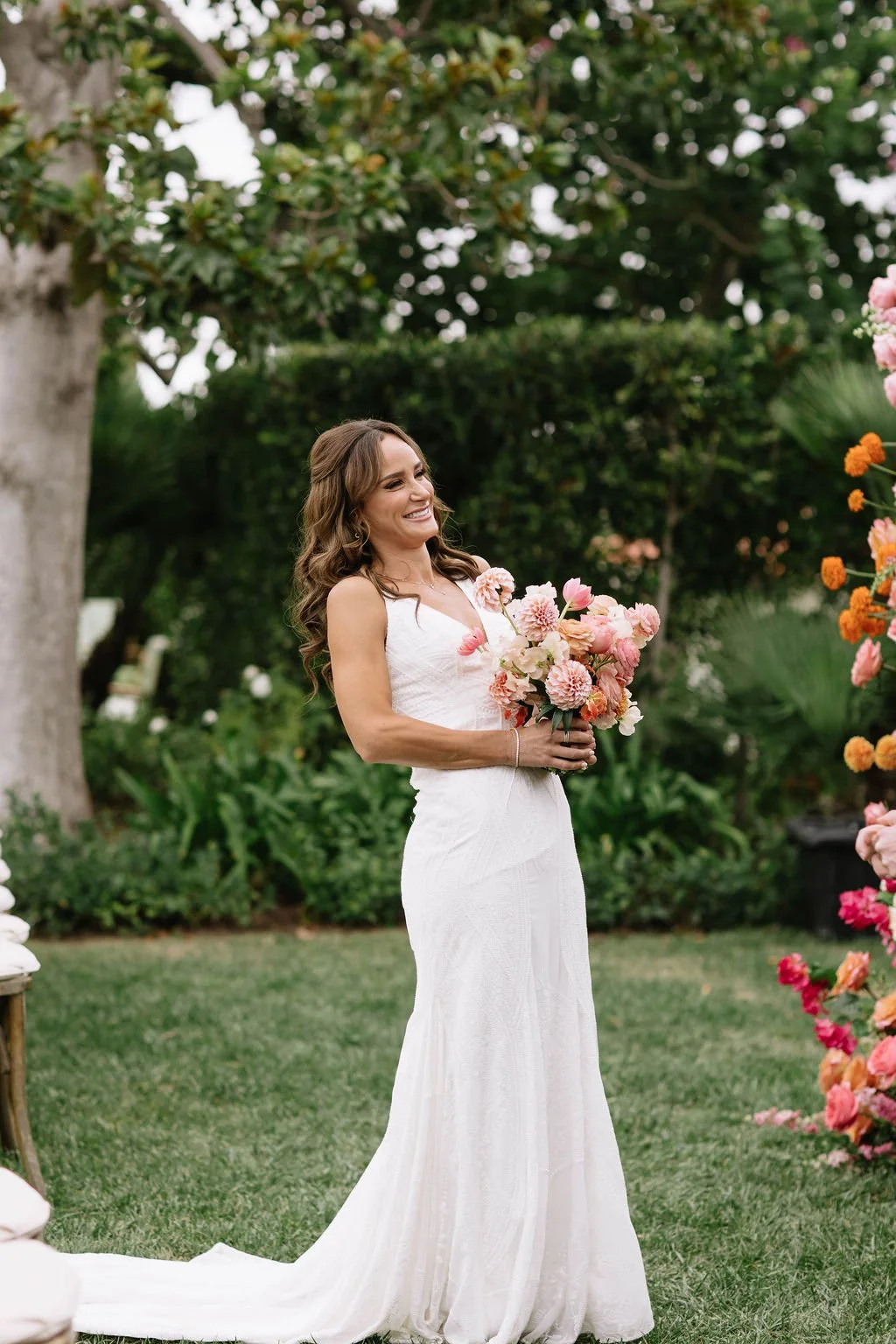 A woman in a white wedding dress holding a bouquet of pink flowers, smiling outdoors amidst greenery and flowers during a wedding celebration.