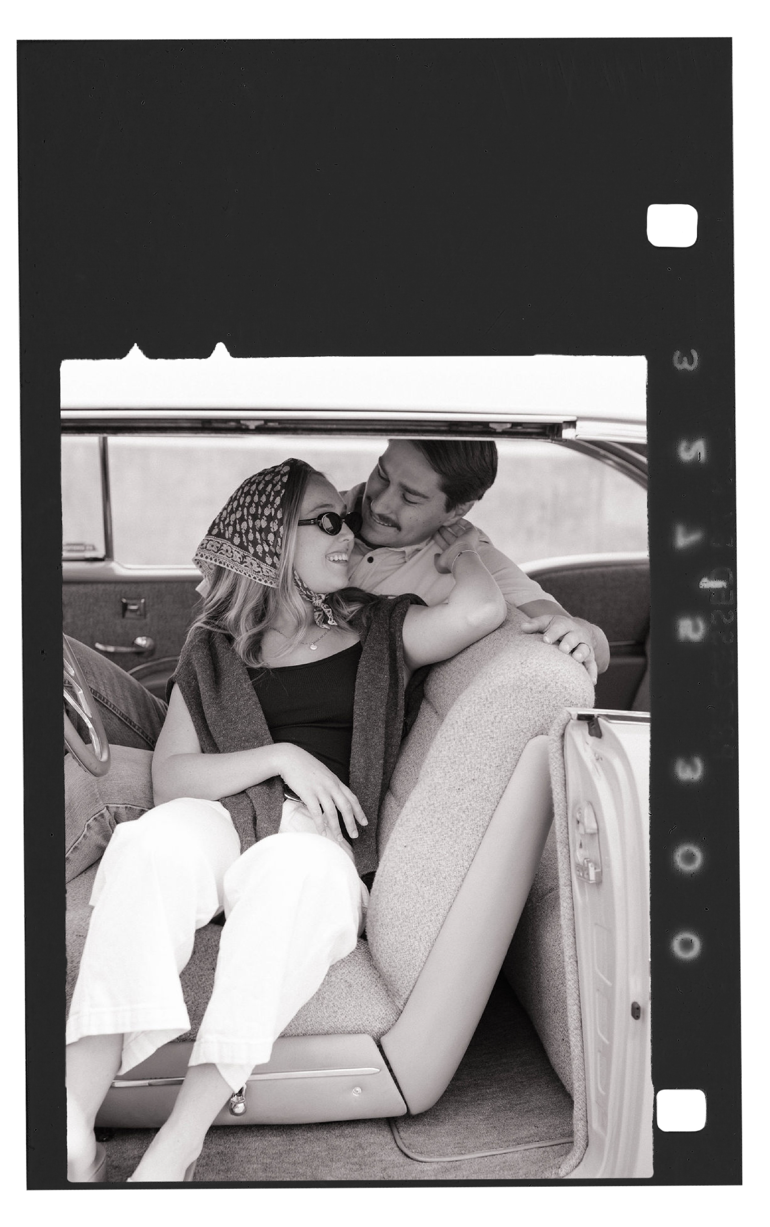 A black and white photo of a man and woman sitting inside an old car, sharing a happy and intimate moment. The woman is wearing sunglasses, a patterned headscarf, and light-colored pants, while the man is smiling and looking at her.