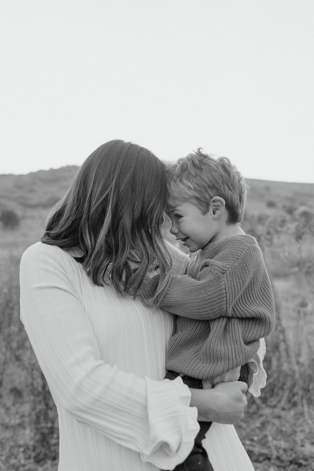 A woman holding a young boy close outdoors, both touching foreheads and smiling in a grassy field with a hill in the background.