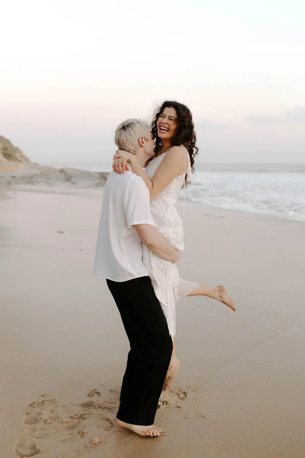 A couple on the beach, the man lifting the woman, both smiling and laughing, during sunset or dawn.