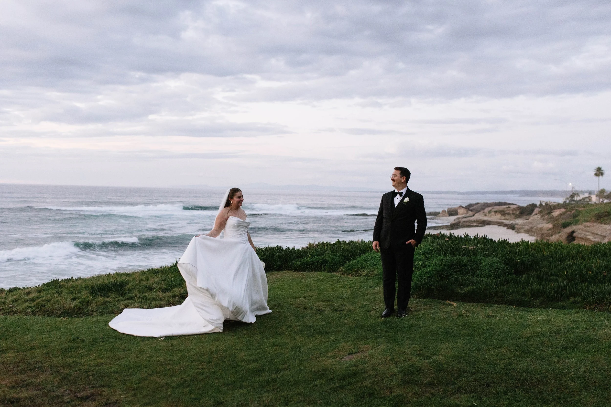 Bride in white wedding dress holding the train, and groom in black tuxedo with bow tie, standing on grassy area by the ocean with rocky shoreline and cloudy sky in the background.