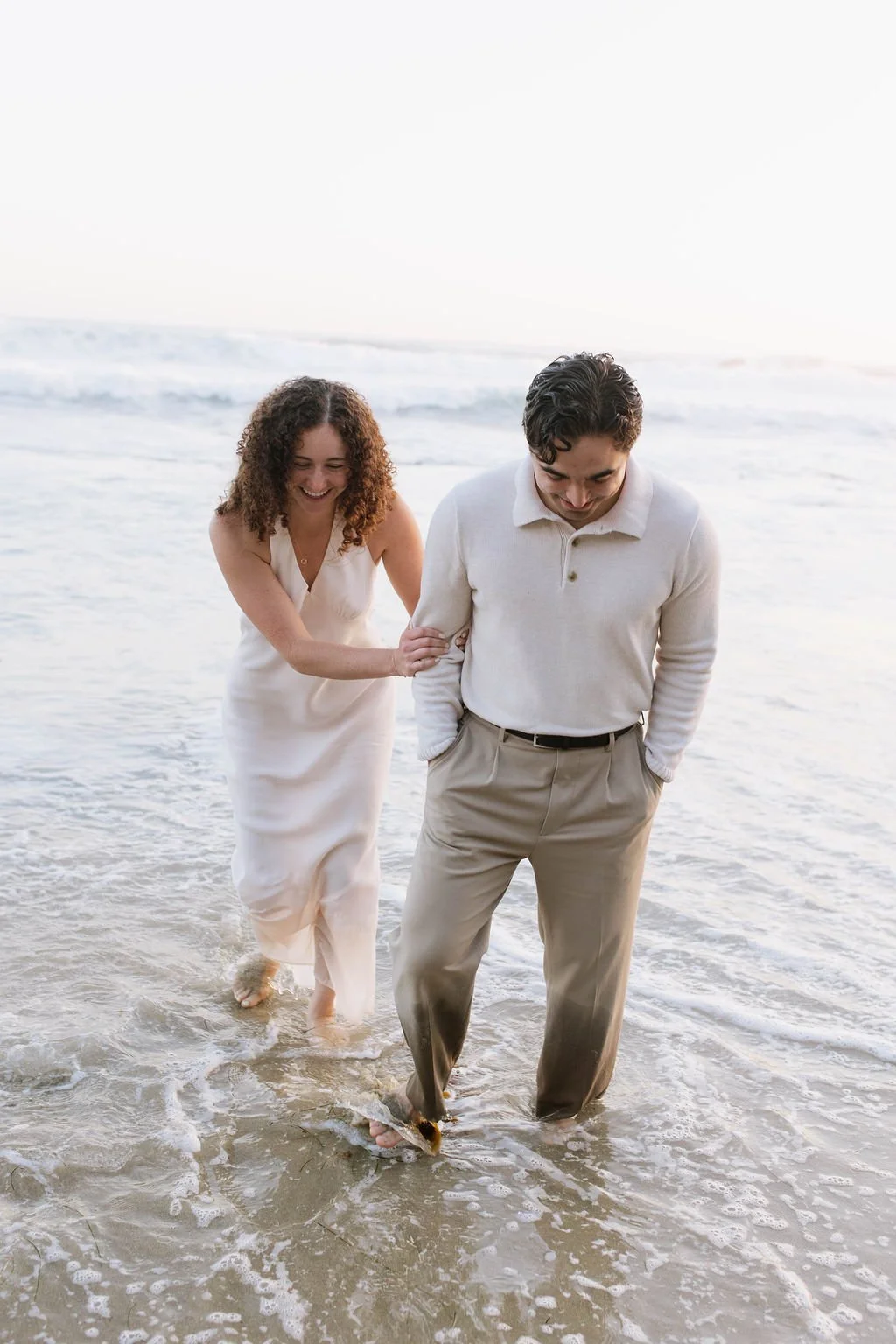 A couple walking in shallow ocean water, smiling, with the woman holding the man's arm, both dressed in light-colored clothing, at the beach during sunset.