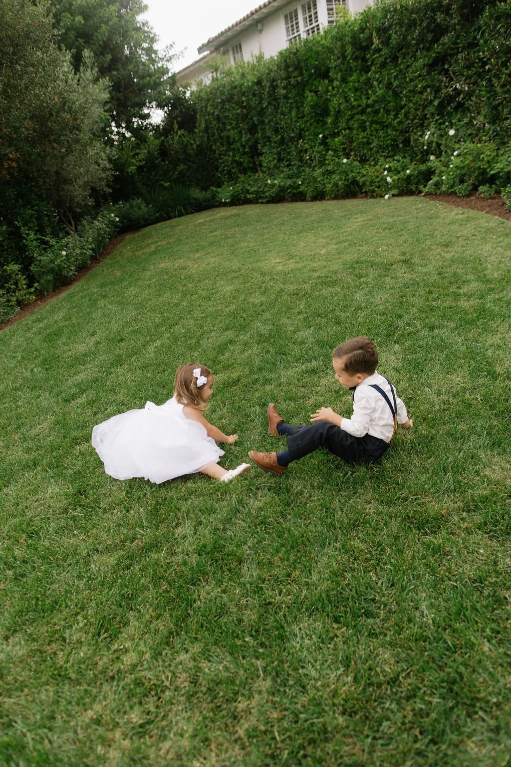 A young boy and girl playing on a lush green lawn in a backyard, with the boy sitting and the girl lying on her stomach, both smiling and engaging with each other.