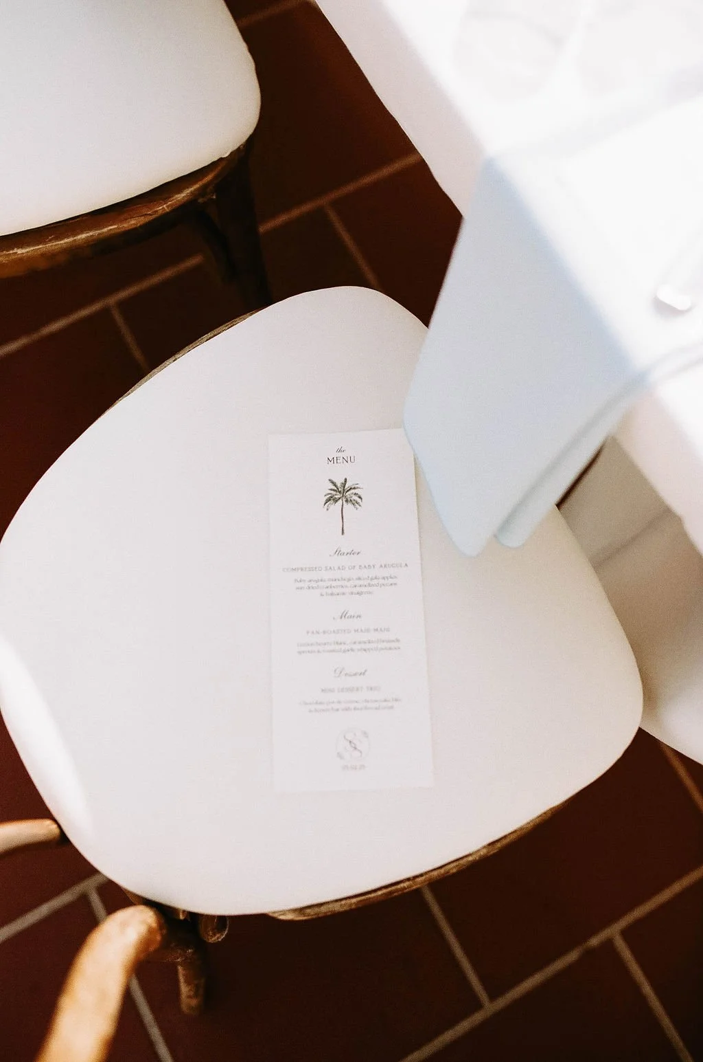 A white chair with a seat covered in a white cloth, holding a small printed menu with a palm tree illustration, placed on the seat. Part of a white tablecloth is visible nearby, and the background shows a reddish-brown tiled floor.