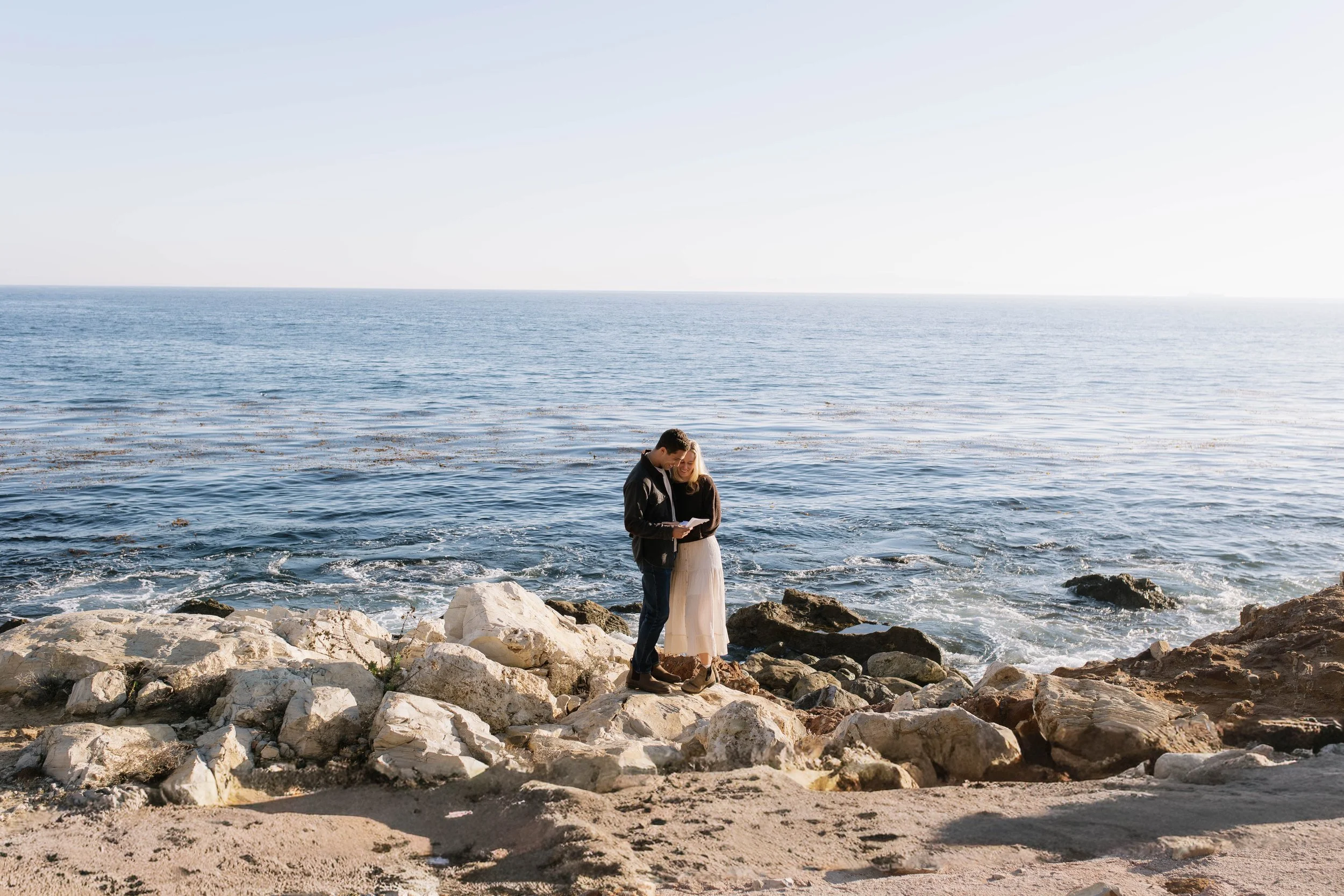 A couple stands on rocks by the ocean, looking at something together during daytime with clear sky.