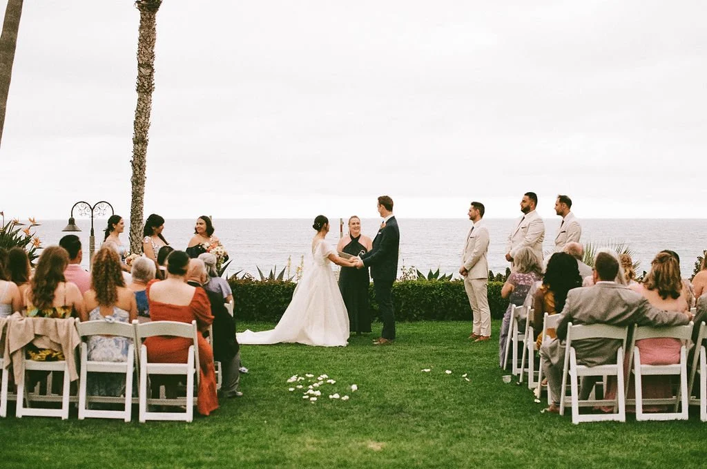A wedding ceremony taking place outdoors by the ocean, with the bride and groom exchanging vows, surrounded by guests seated on white chairs, with a palm tree and ocean in the background.