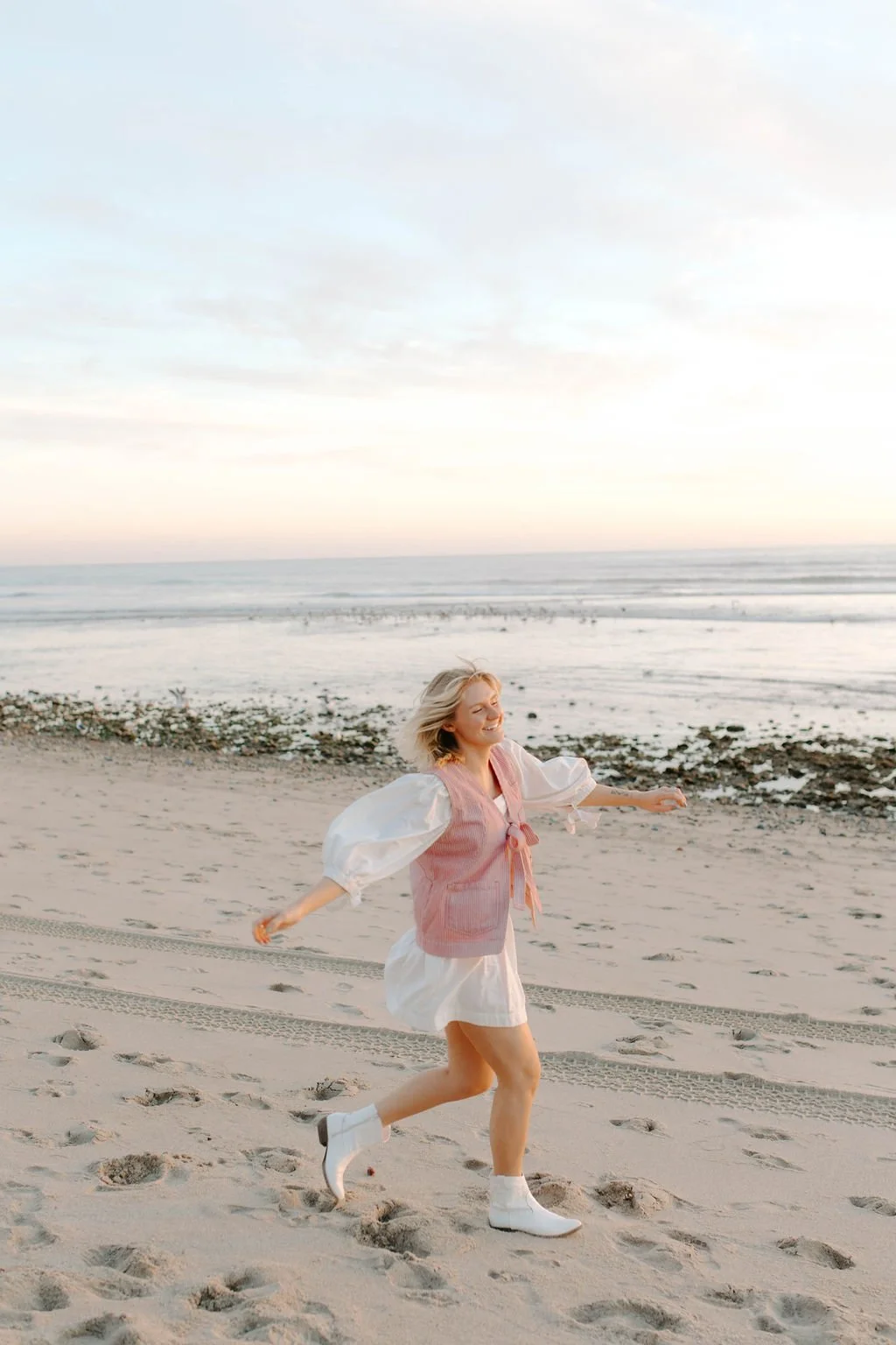 A woman with blonde hair running and smiling on the beach during sunset, wearing white boots, a white dress, and a pink vest.