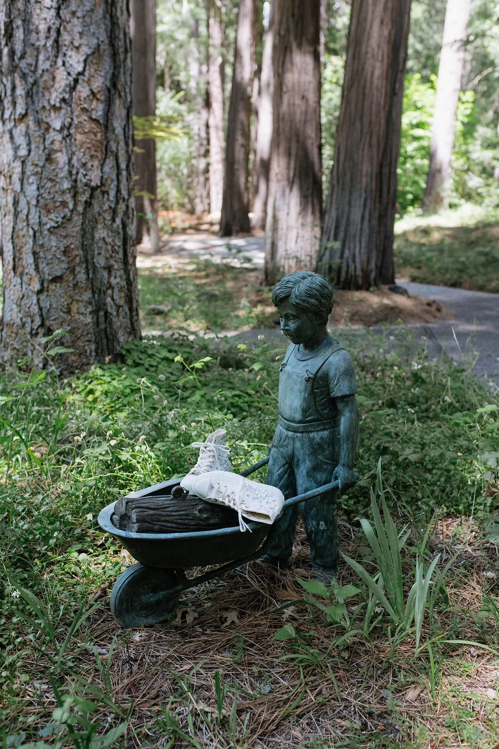 A bronze statue of a young boy pushing a wheelbarrow filled with a pair of sneakers and a piece of wood, situated outdoors among trees and greenery.