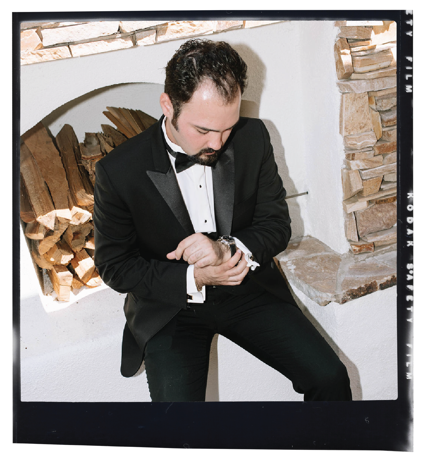 A man in a black tuxedo checking his watch in front of a stone fireplace.